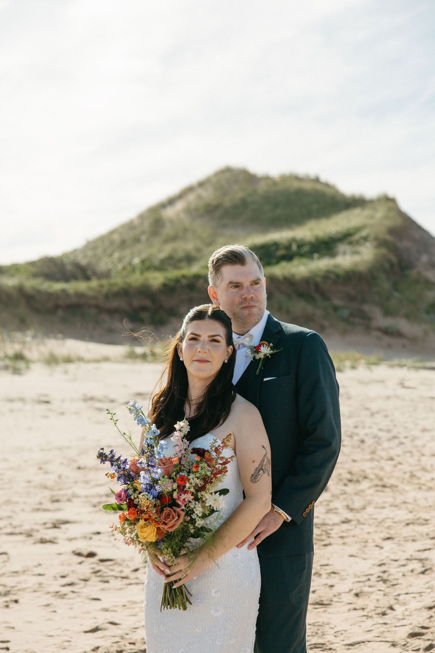 Bride holding a vibrant wildflower bouquet is embraced by her groom on a sandy beach with grassy dunes behind them.