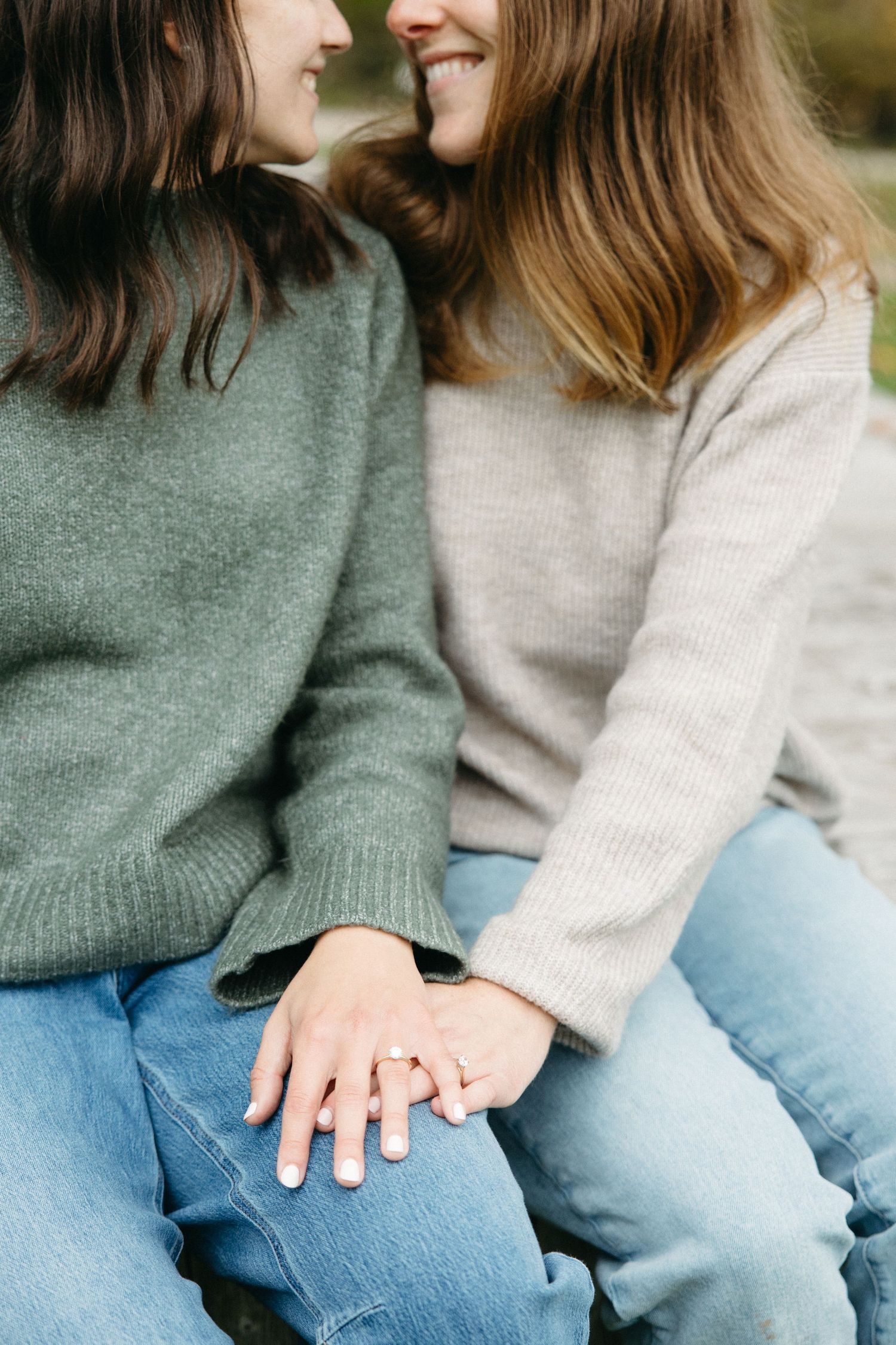 Two women in cozy sweaters and jeans sit close together, smiling and touching foreheads as their hands rest together, one wearing a ring.