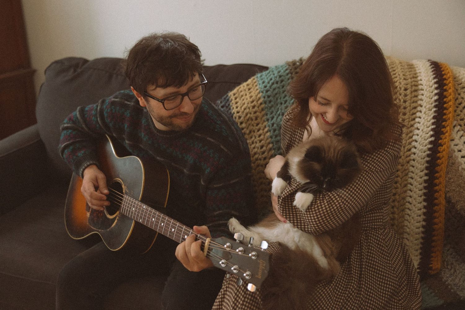 Man playing acoustic guitar on sofa while woman holds a cat, cozy home setting with colorful blanket.