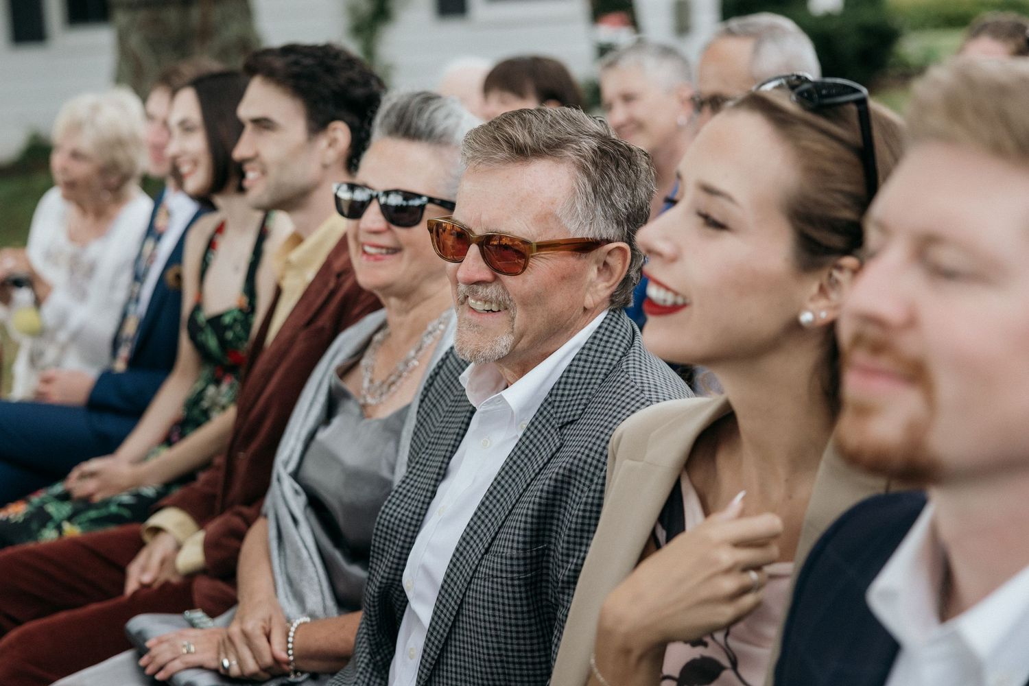 Wedding guests seated outdoors smile and laugh during ceremony, several wearing sunglasses on sunny day.