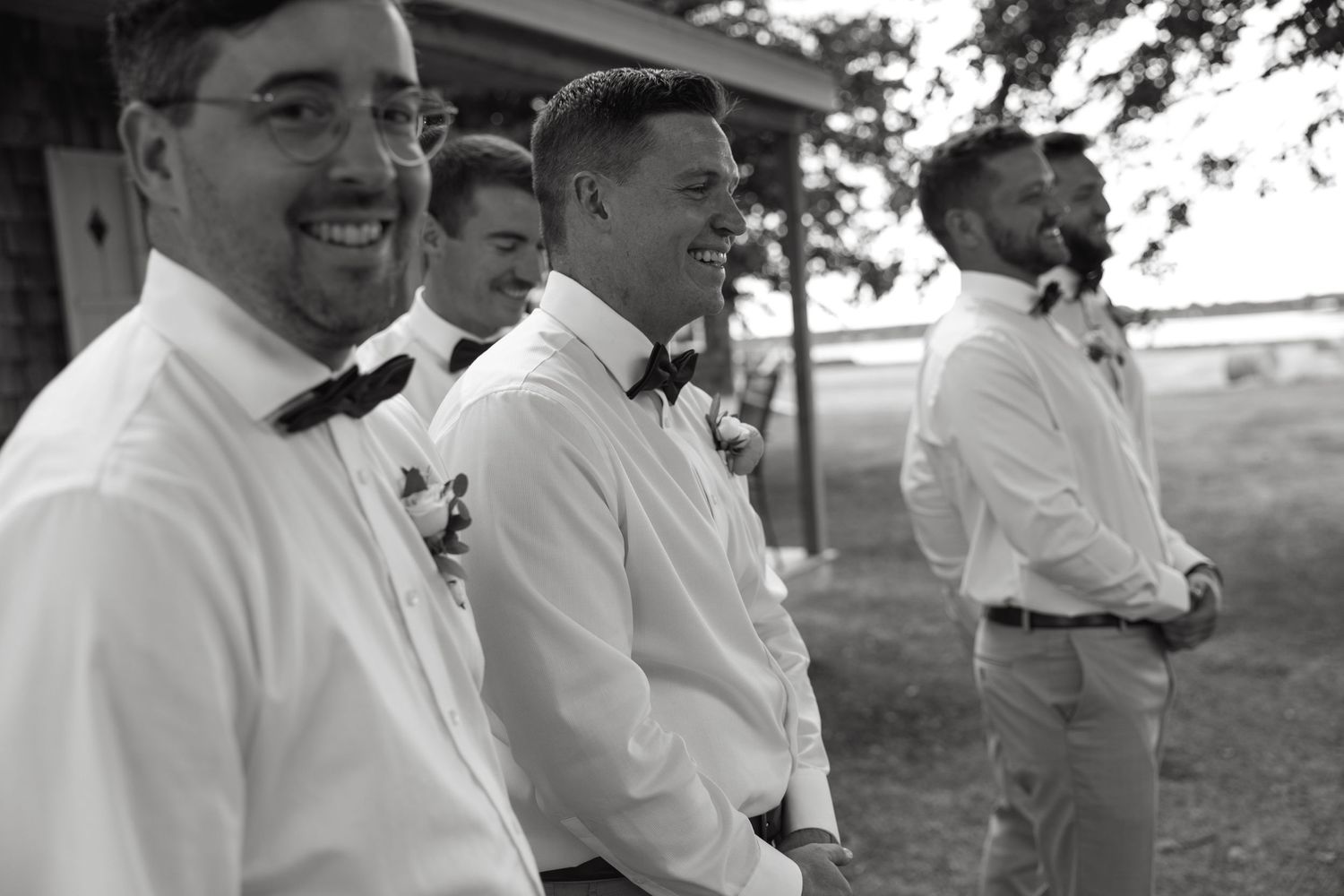 Groomsmen in white shirts and bow ties laugh together during an outdoor wedding ceremony.
