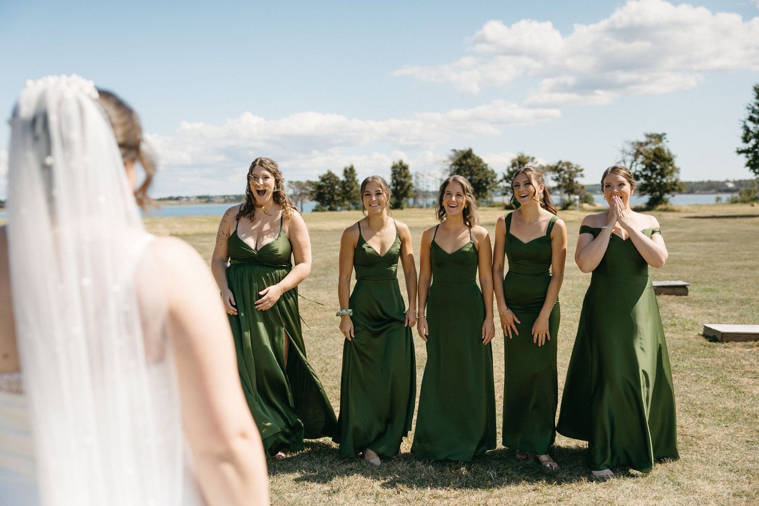 Bride in white gown and veil shares first look moment with five bridesmaids in emerald green dresses outdoors.
