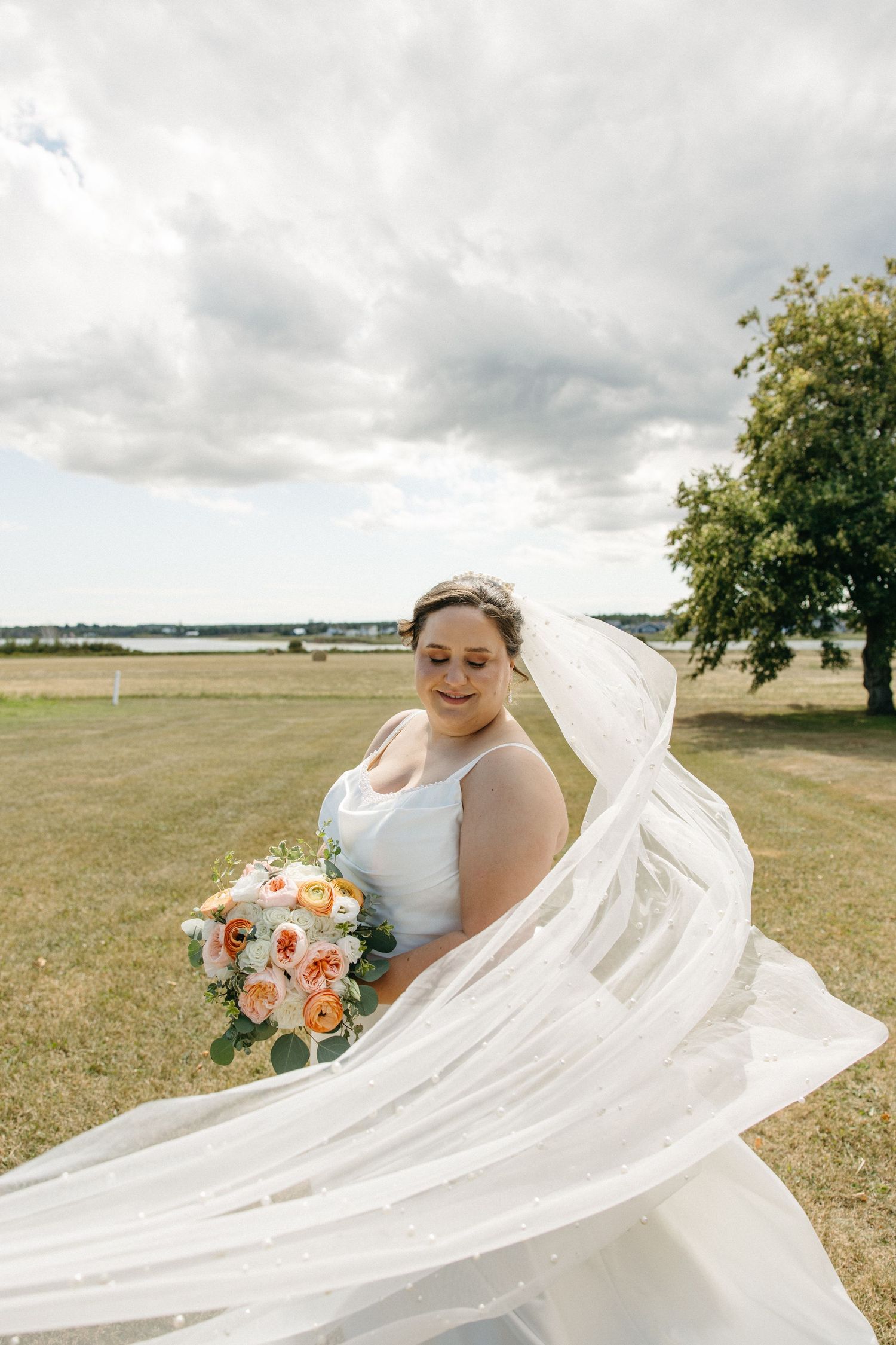 Bride in white gown with flowing veil holding peach bouquet outdoors.