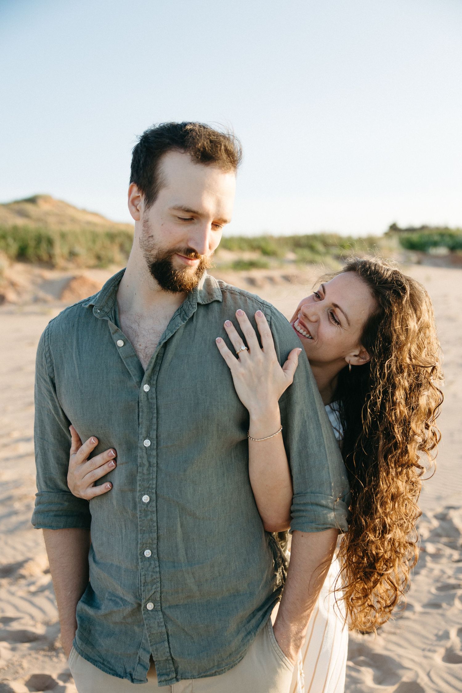 A bearded man and curly-haired woman share a joyful moment on a sandy beach, she smiling and resting her hand on his chest.