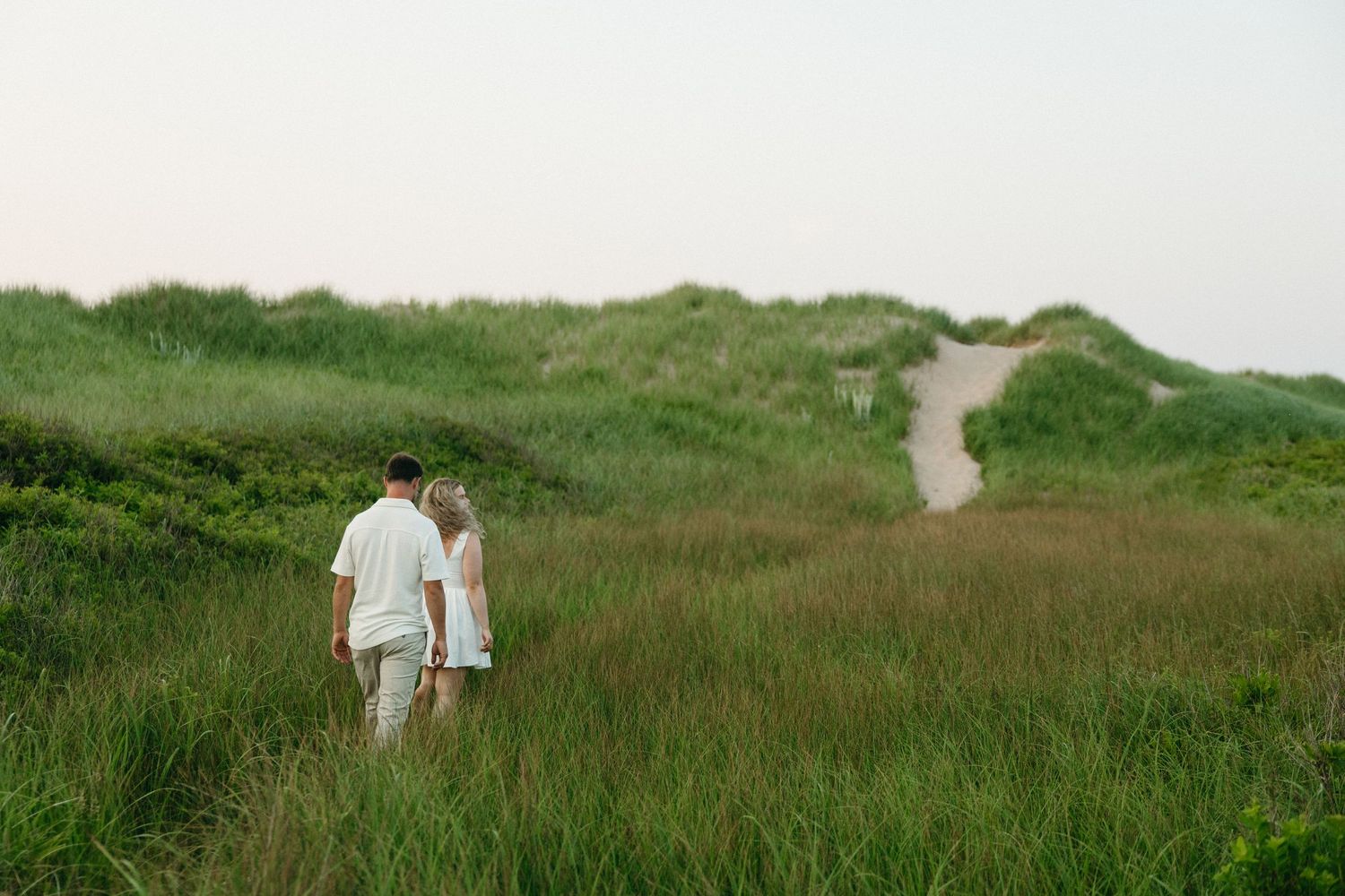 Couple walking hand in hand through lush green coastal dunes on a serene, overcast day.