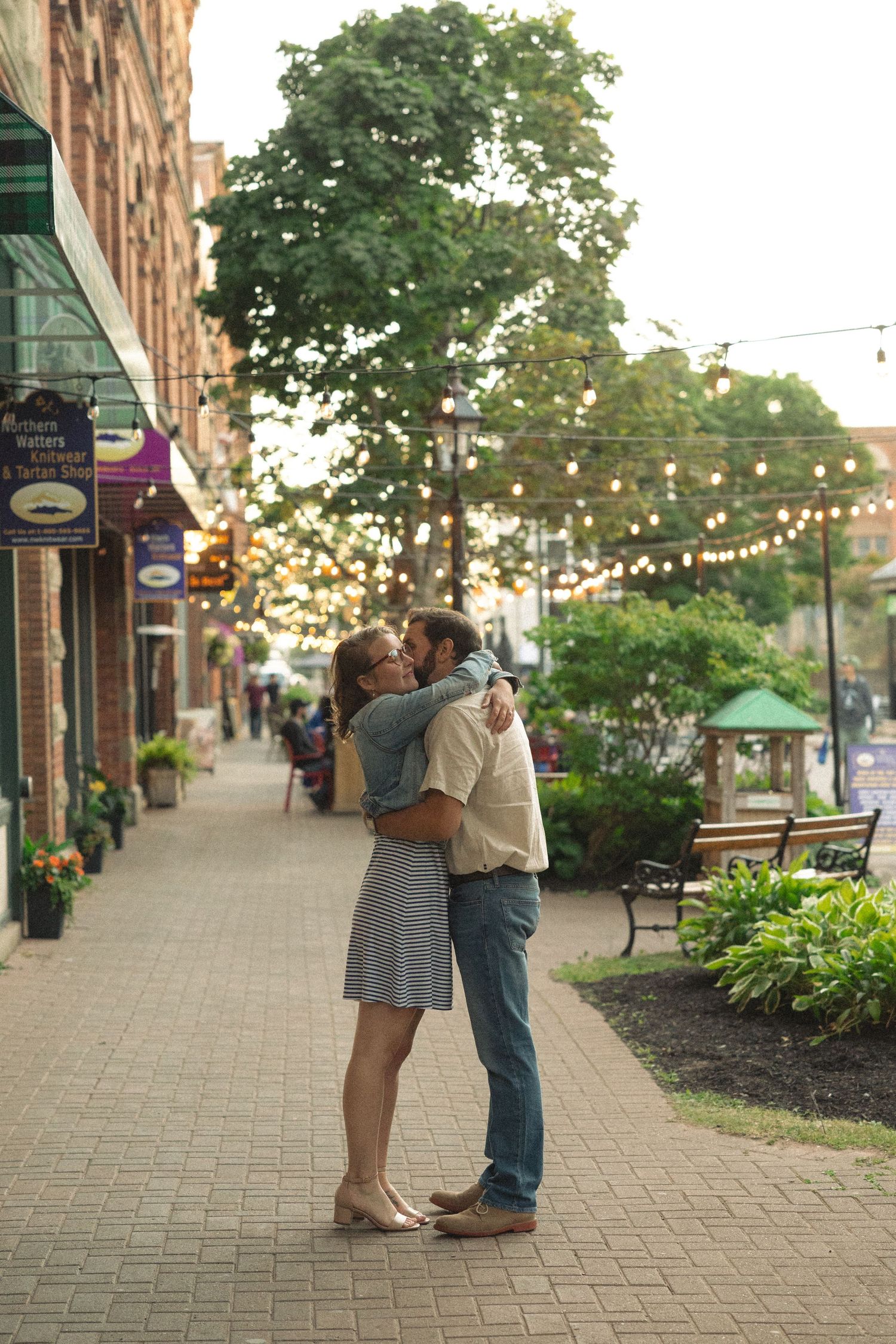 Romantic evening scene on brick-paved street with string lights and storefronts.