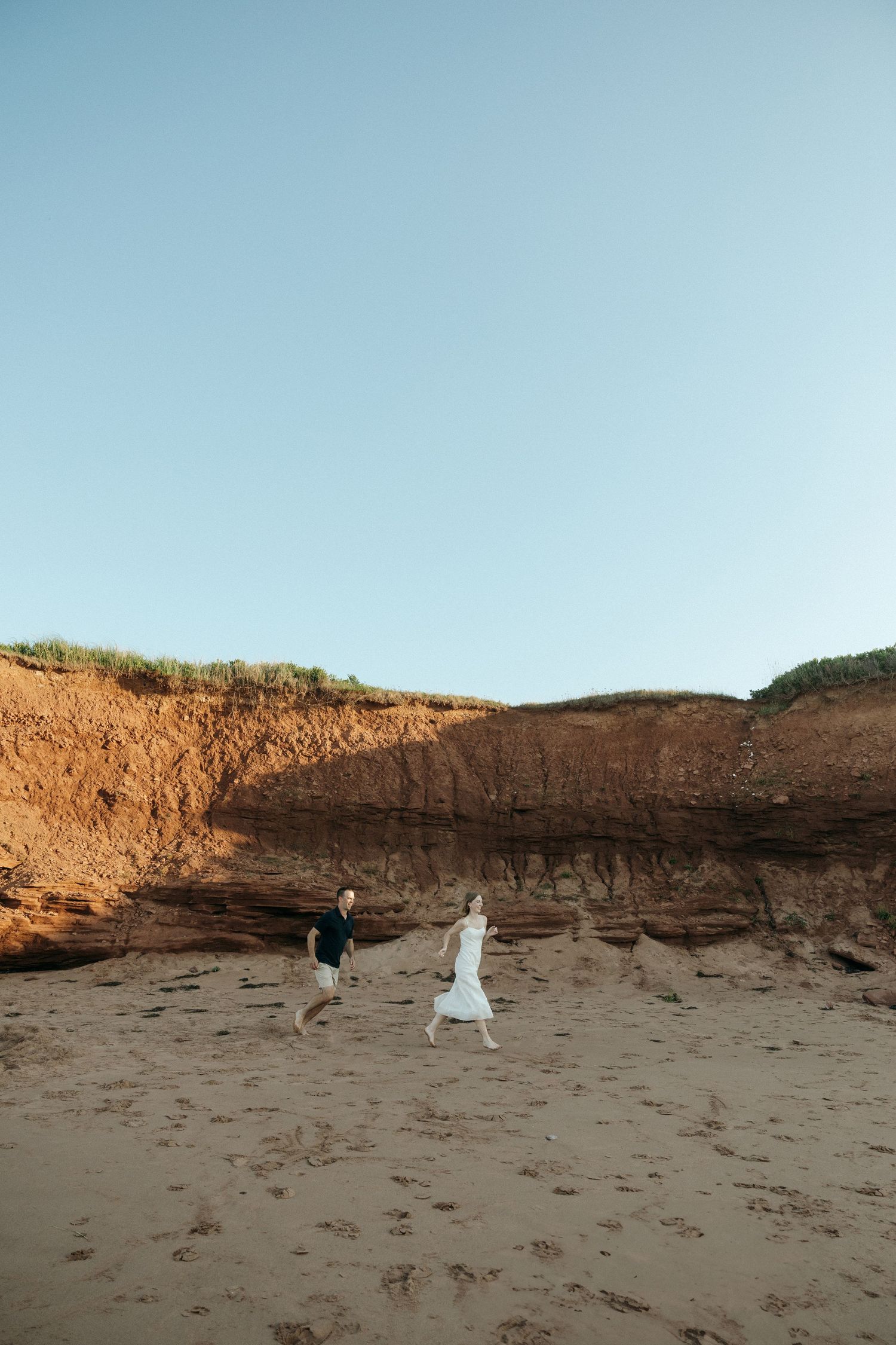 Wide shot of couple walking on beach with dramatic cliff backdrop.
