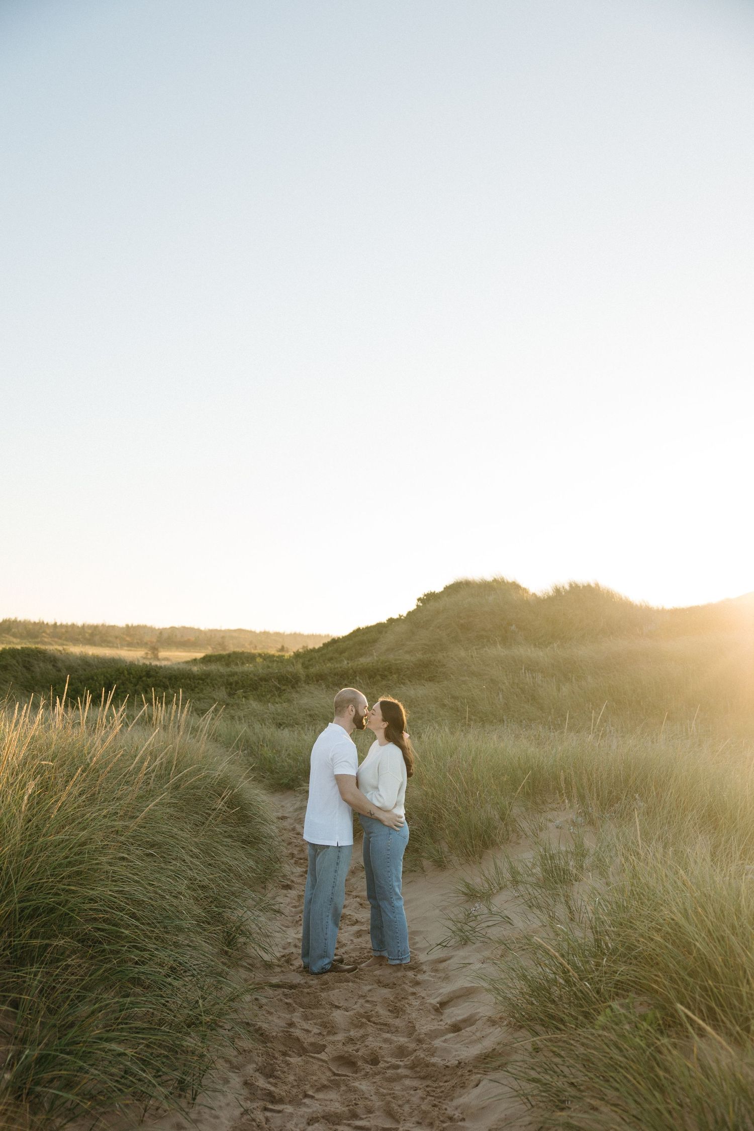 Two people share intimate moment on sandy path through beach grass at dusk.