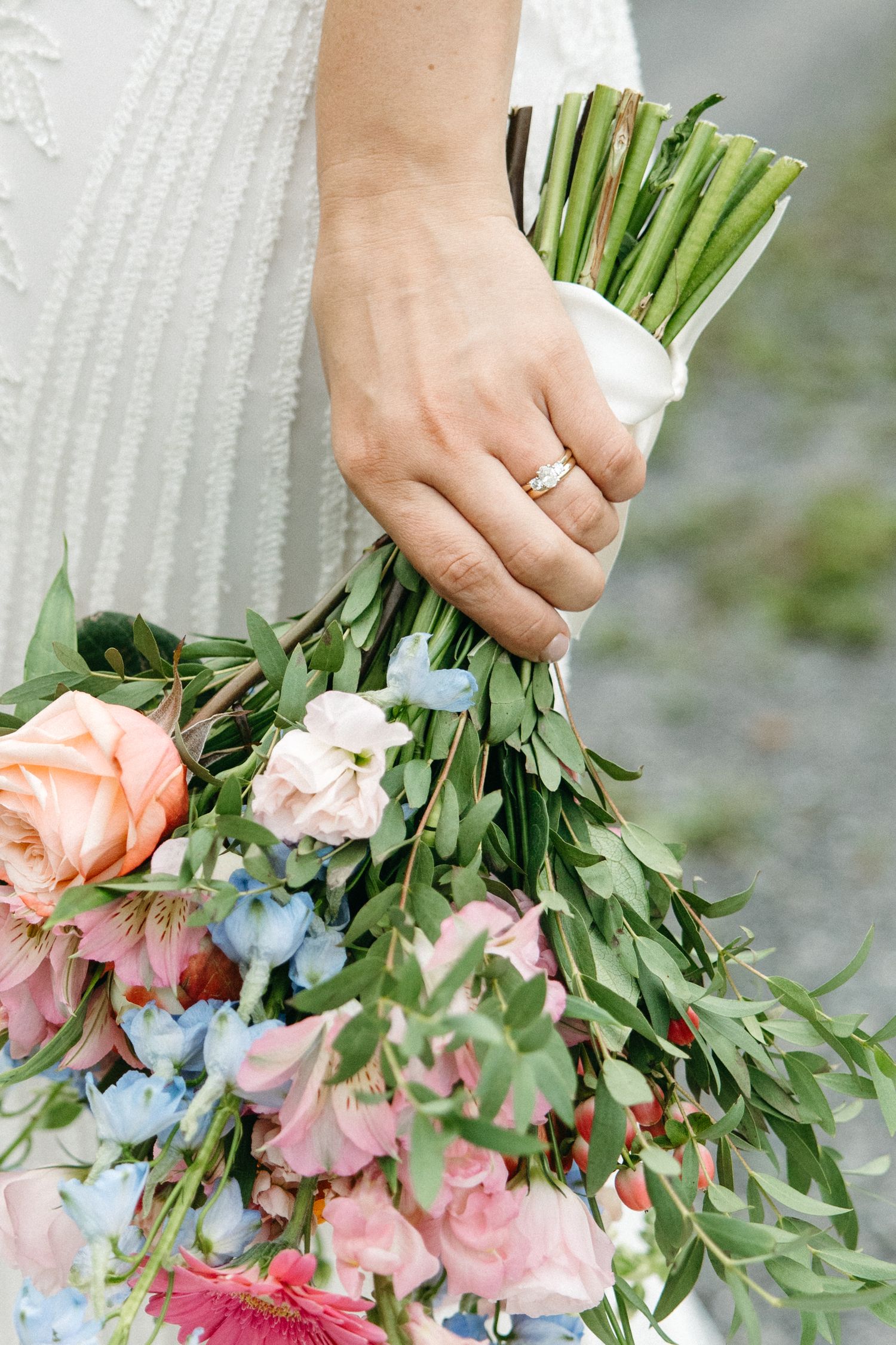 Close-up of bridal bouquet with pink roses and blue flowers, showing wedding ring detail.