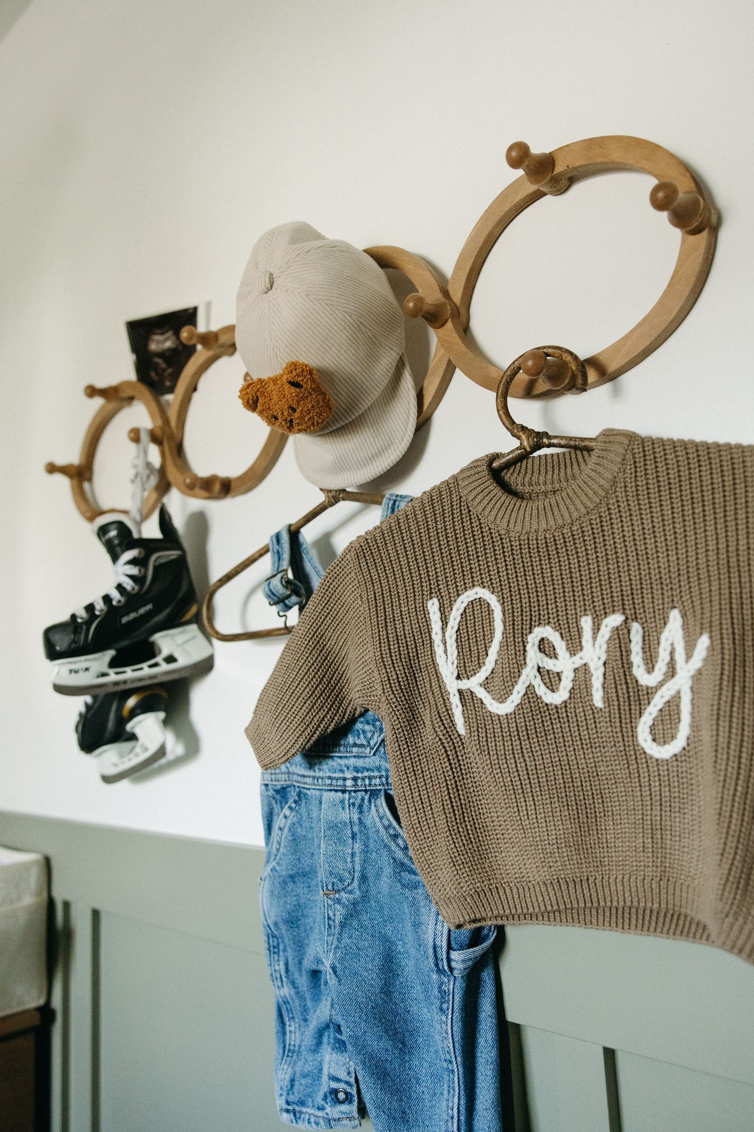 A brown knit sweater with 'Kory' text and denim overalls hanging on wooden wall hooks.