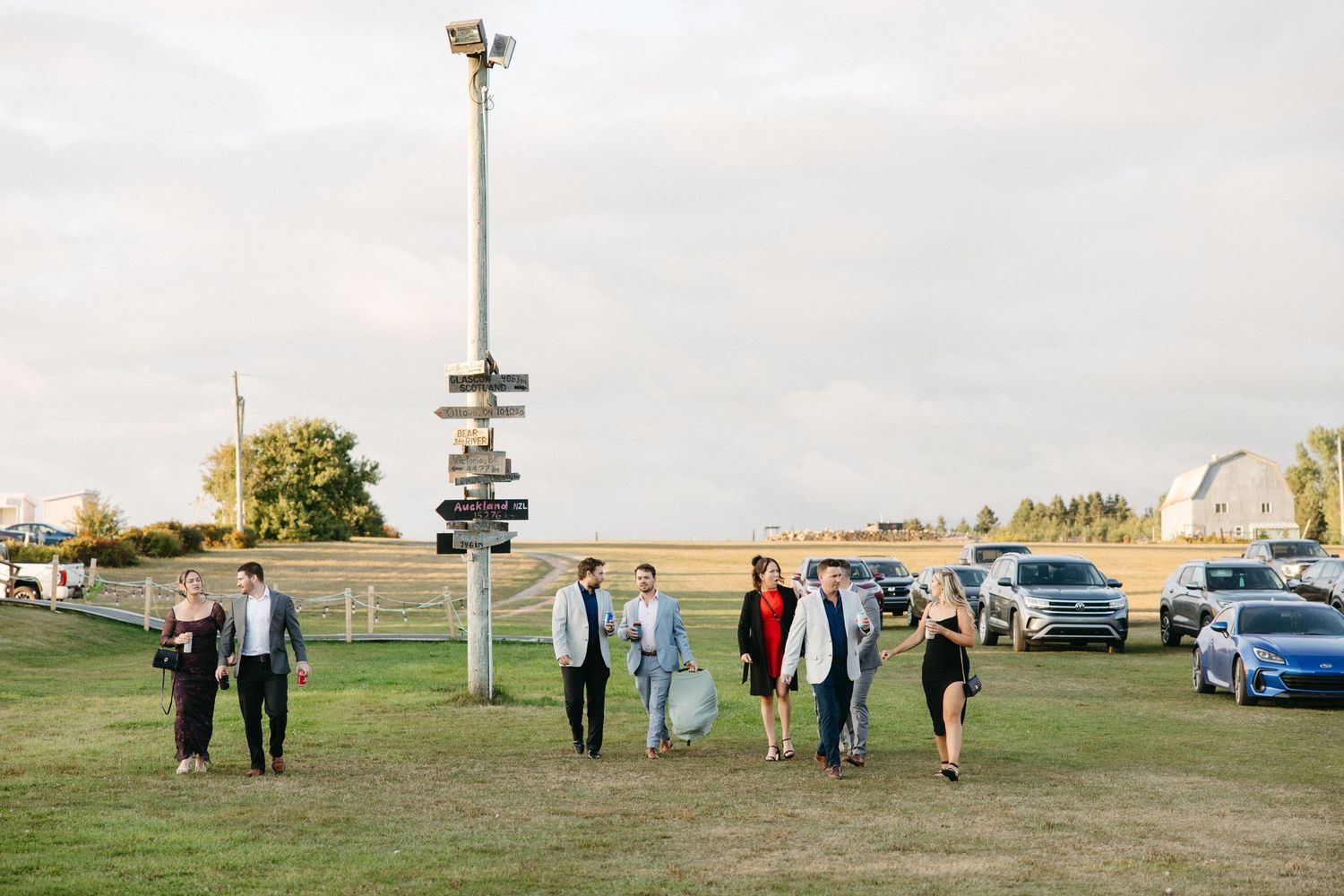 Wide shot of wedding guests scattered across an open field near parked cars.