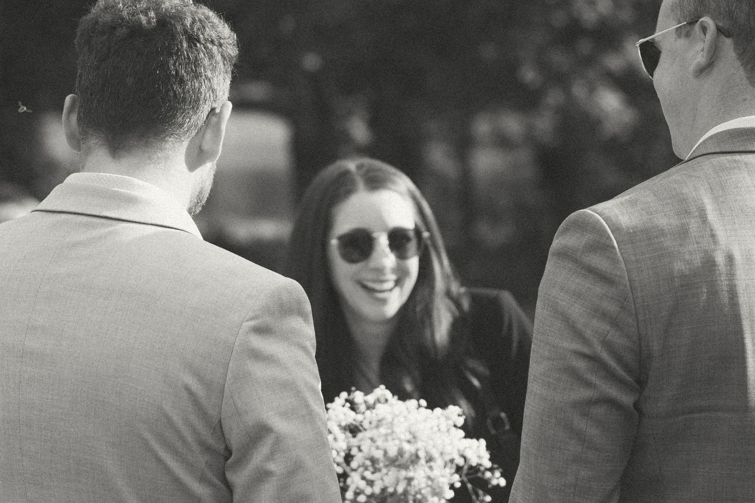 Wedding guests gather outdoors on a sunny day for a celebration under tall trees.