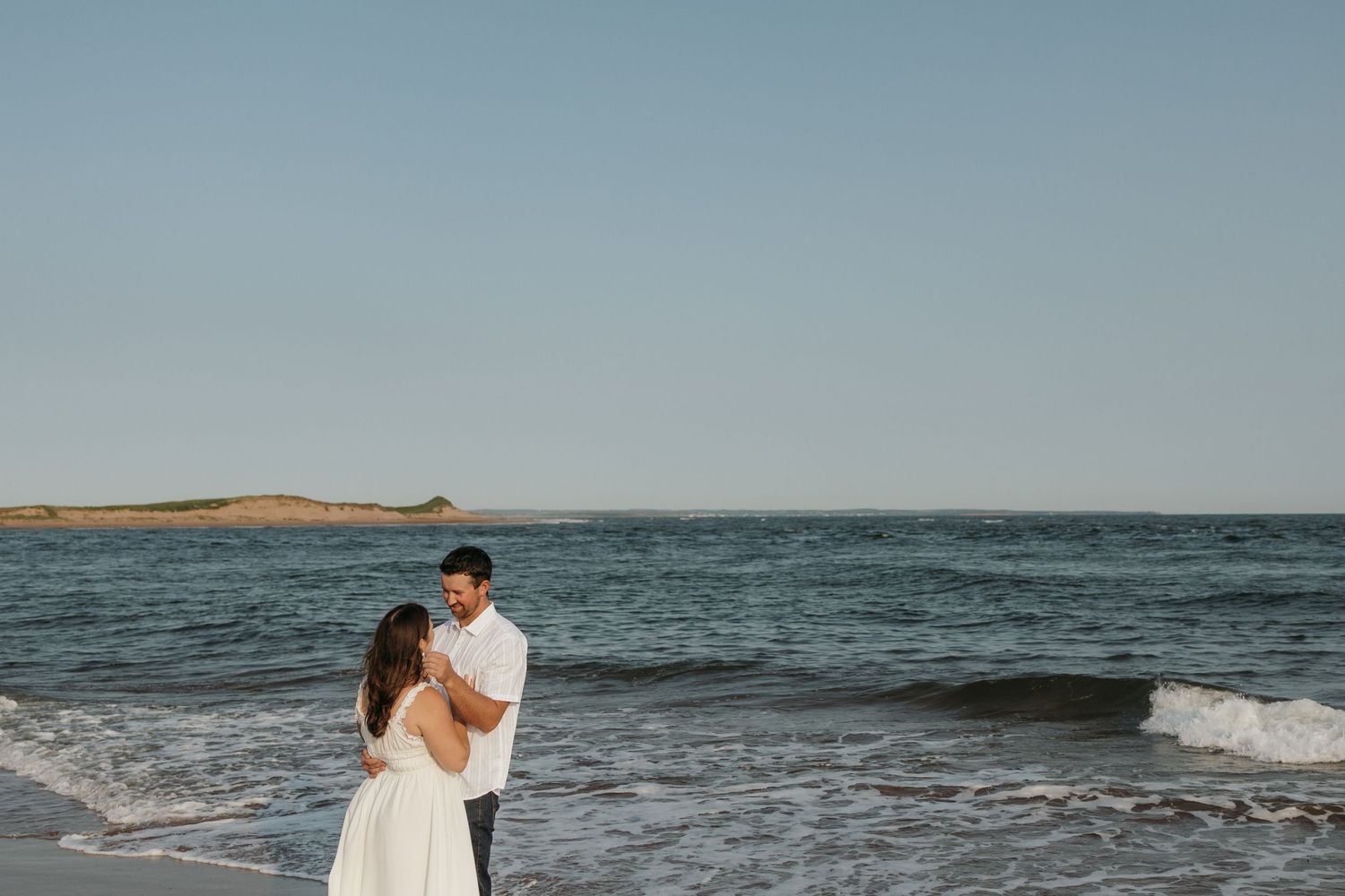 Couple embracing on a beach with blue ocean waves and clear sky in background.