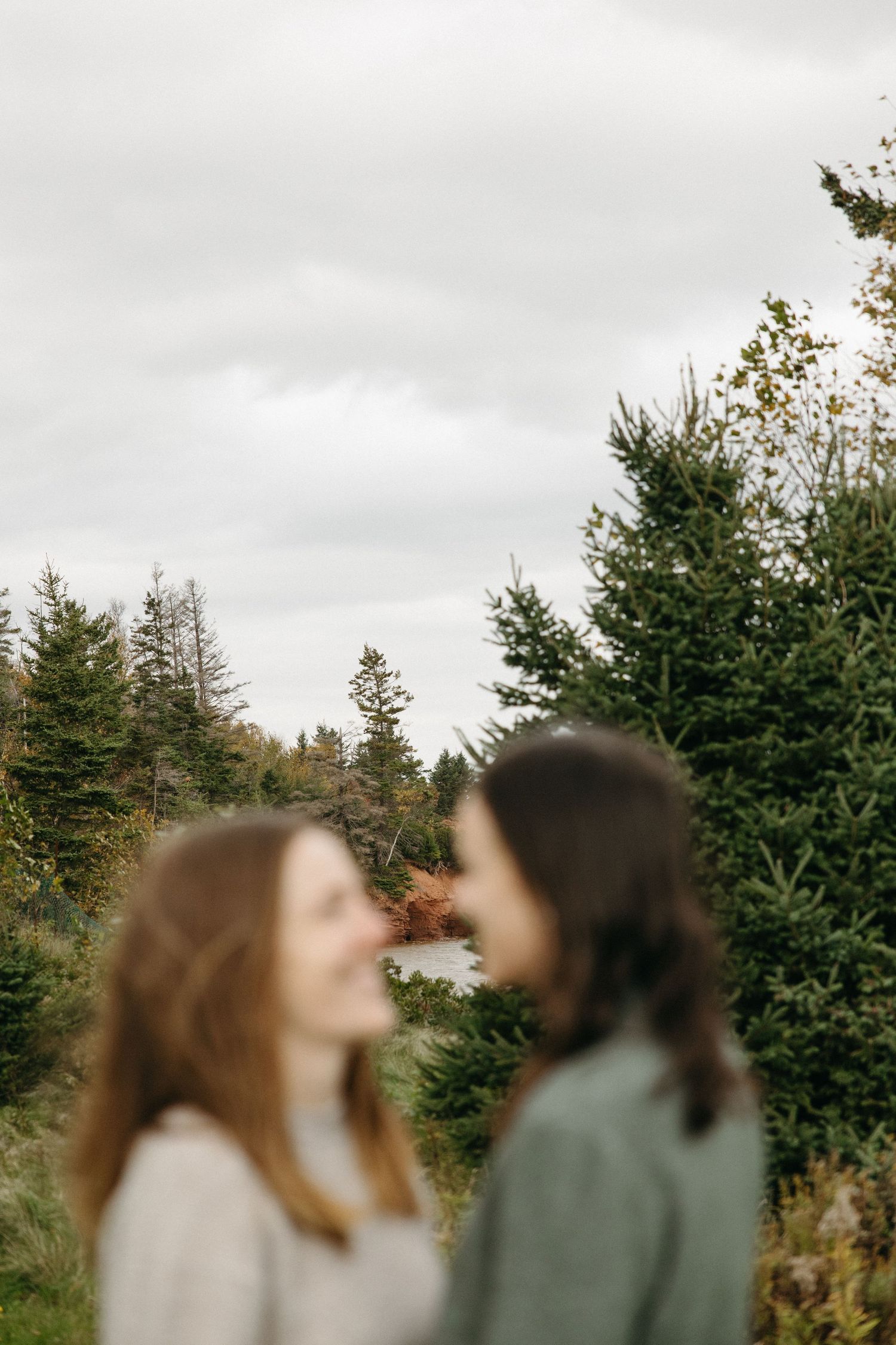 Two women with long hair smile at each other in a blurred foreground, with evergreen trees and a calm pond under a cloudy sky behind them.