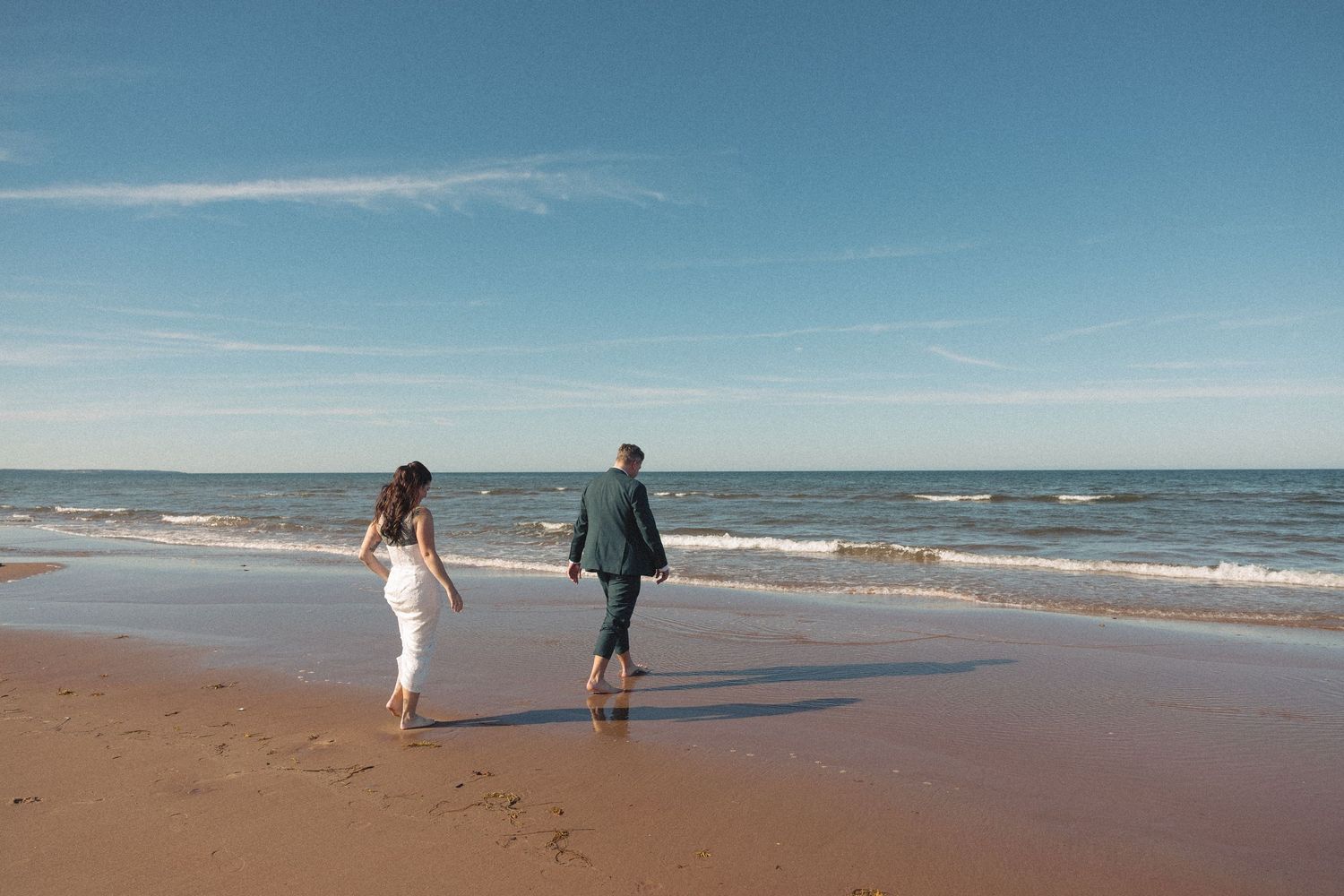 Couple in wedding attire walking barefoot along a sandy beach toward gentle ocean waves under a bright blue sky.