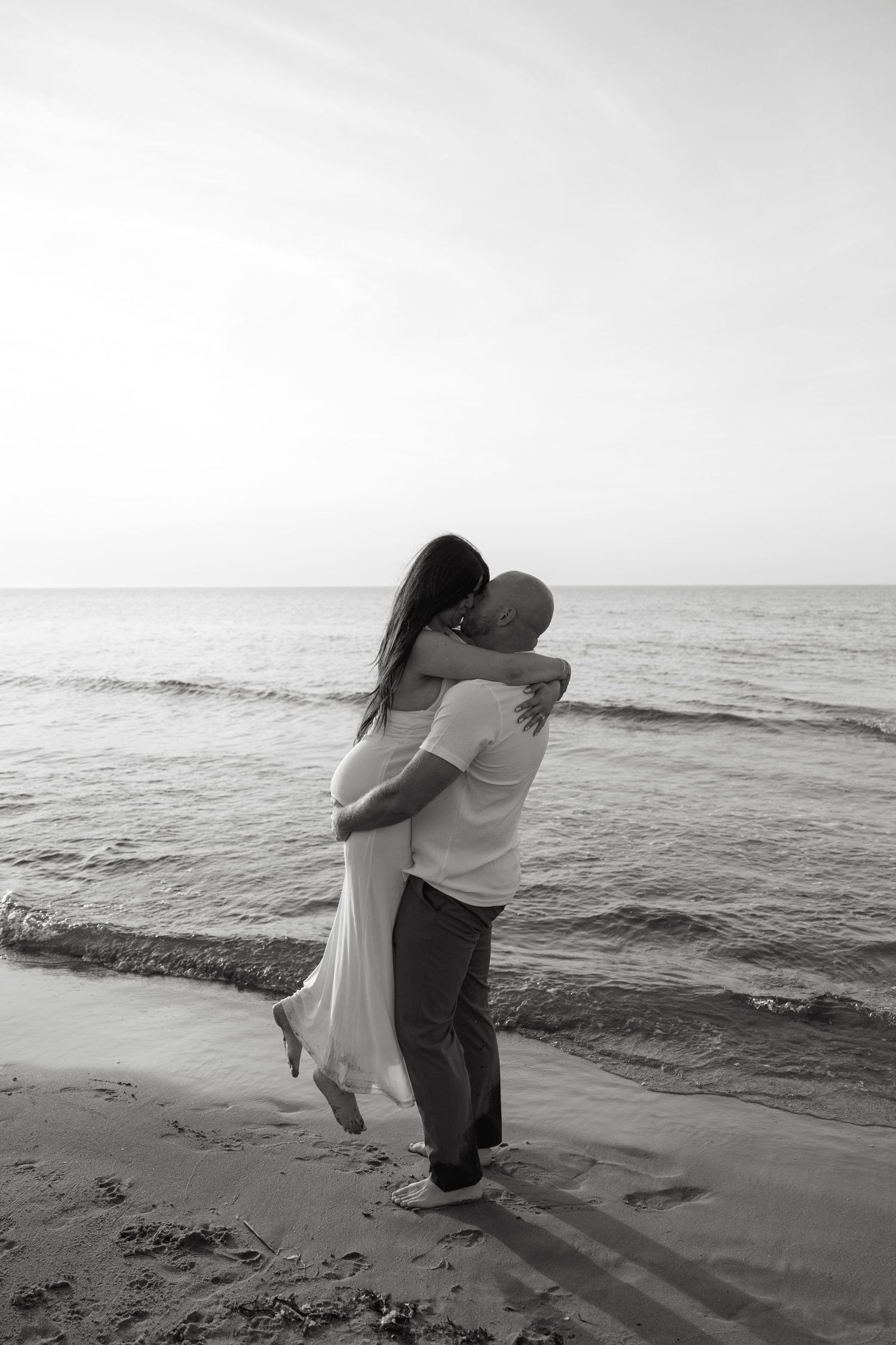 Black and white image of man lifting woman in romantic beach embrace at ocean shoreline.