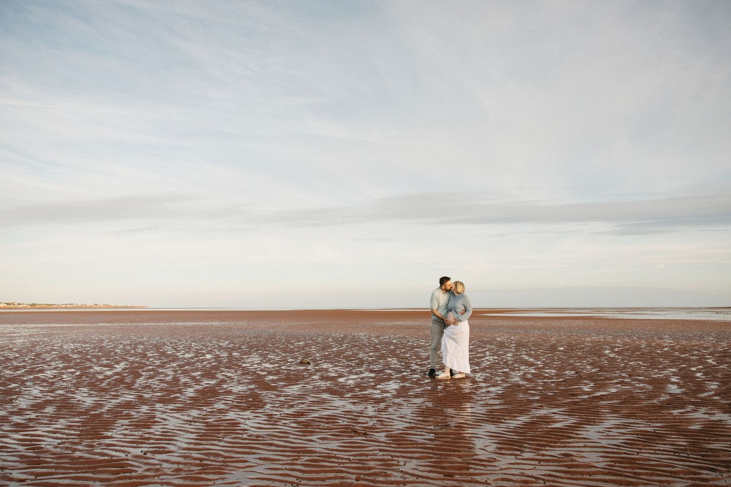 Couple embracing on vast sandy beach shoreline during golden hour maternity photography session.