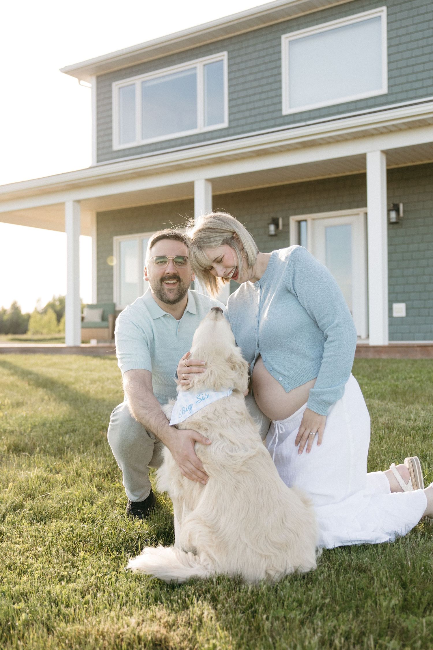 Joyful expecting couple petting their golden retriever on lawn outside their green two-story house.