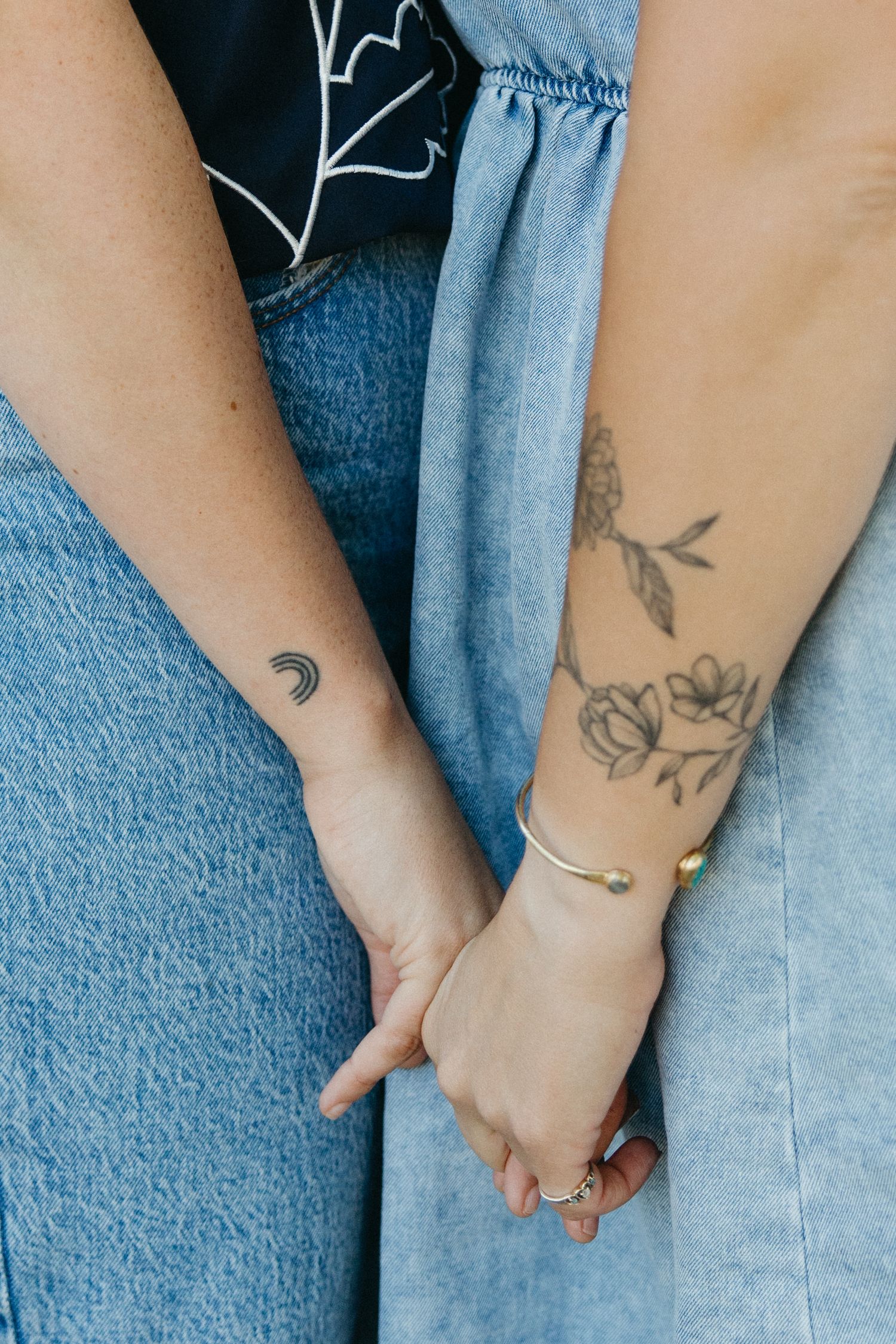 Close-up of hands with delicate floral tattoos and a crescent moon tattoo wearing denim and jewelry.