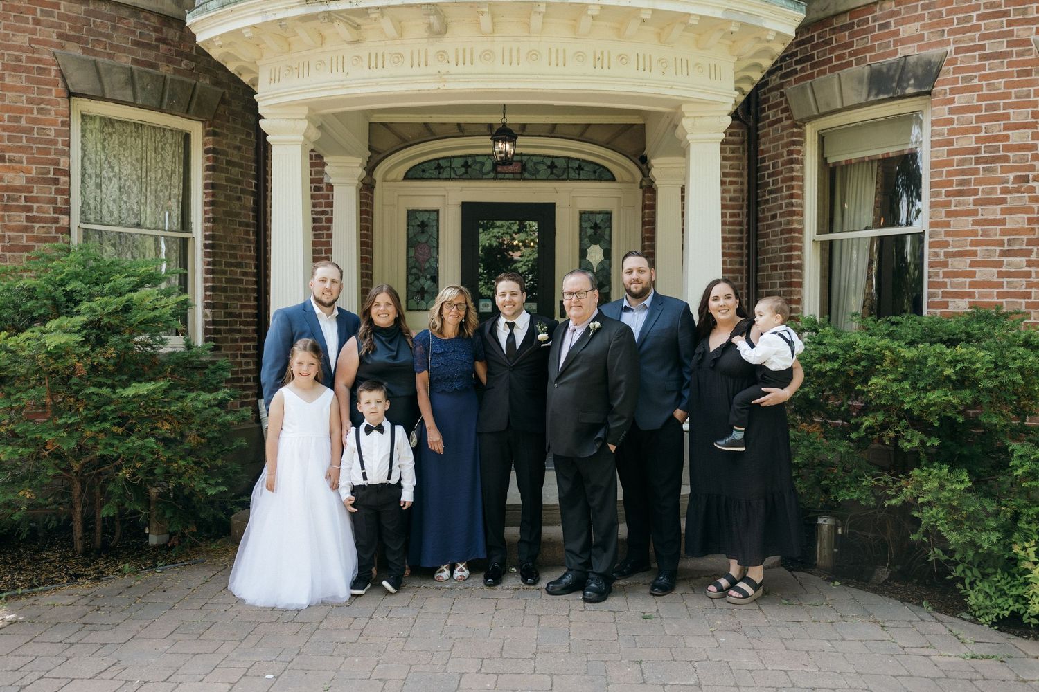 Wedding party and guests posed on the front steps of an elegant brick mansion with white columns.