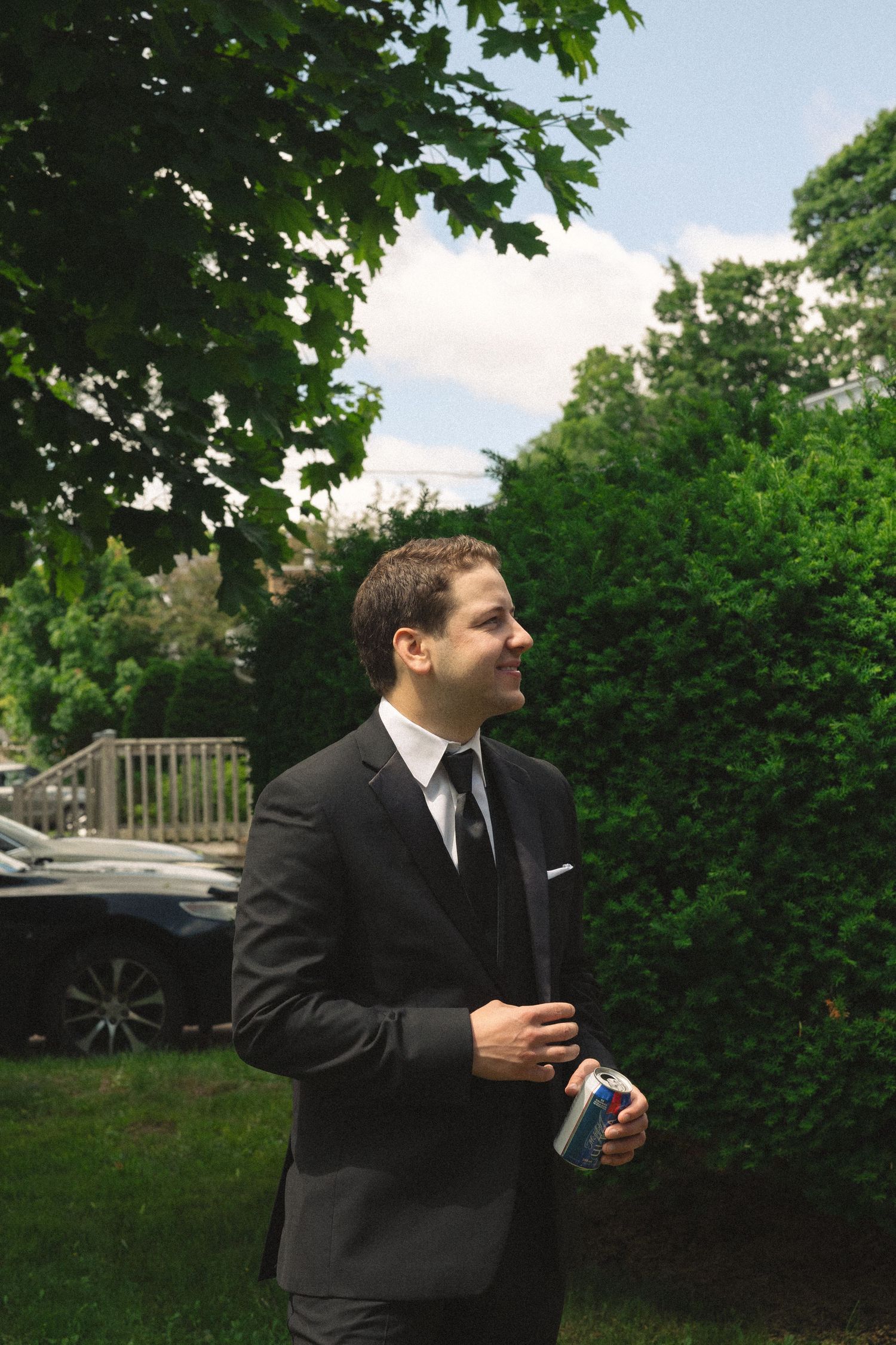 Groomsmen in black suits and ties gather outdoors near green foliage.