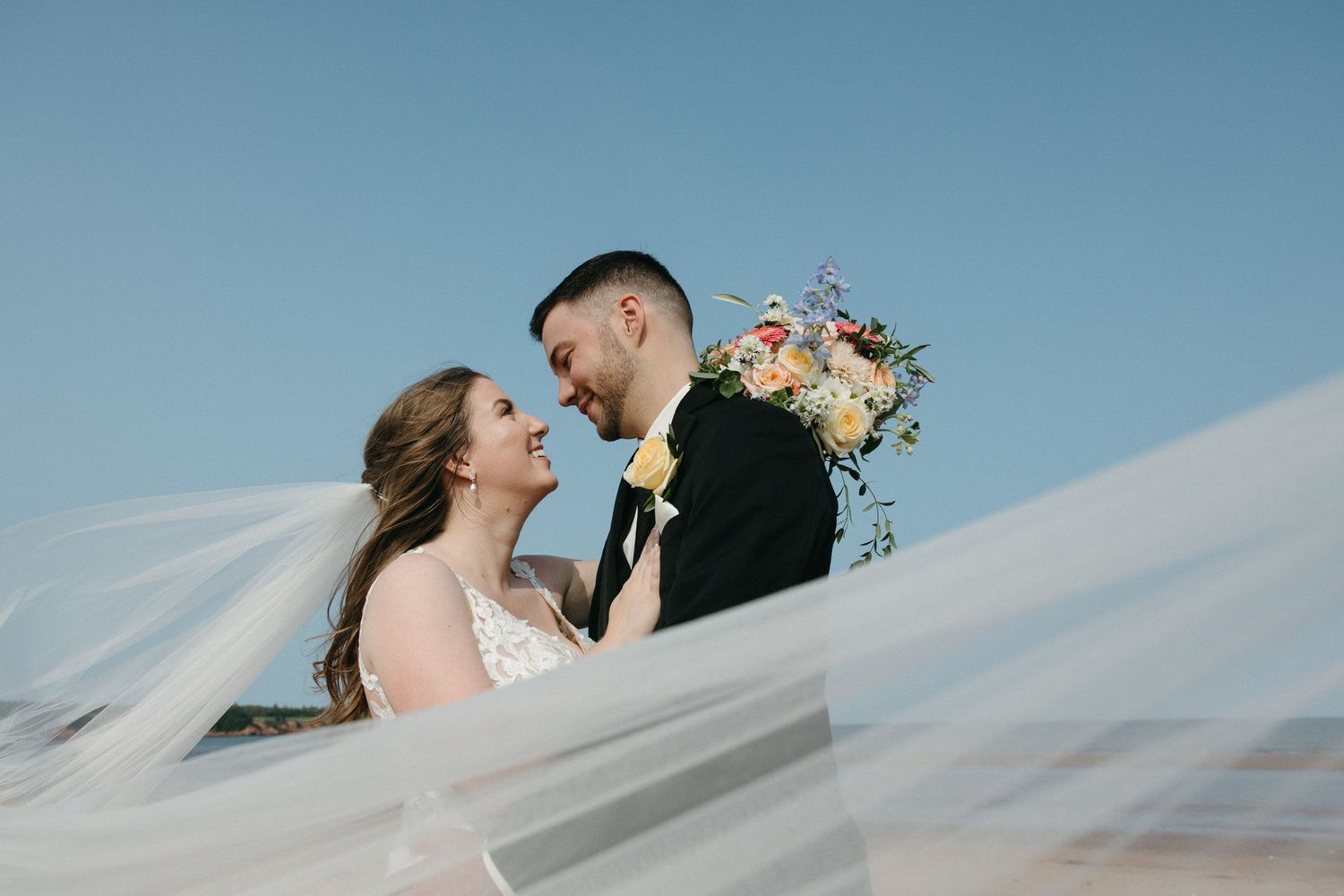 Romantic beach wedding portraits with flowing veil against blue sky.