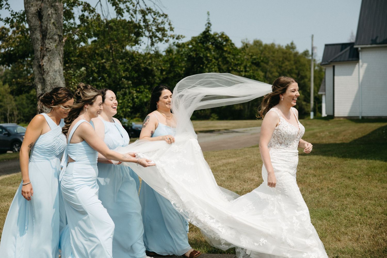 A group in light blue dresses and a white gown walk together on a sunny outdoor path.