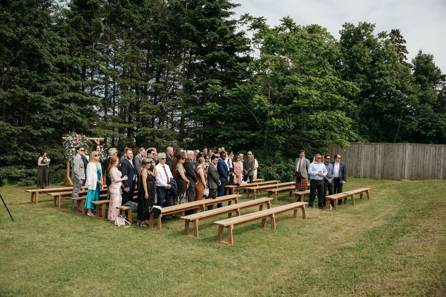 Panoramic view of wooden benches set up for outdoor wedding ceremony with evergreen trees in background.
