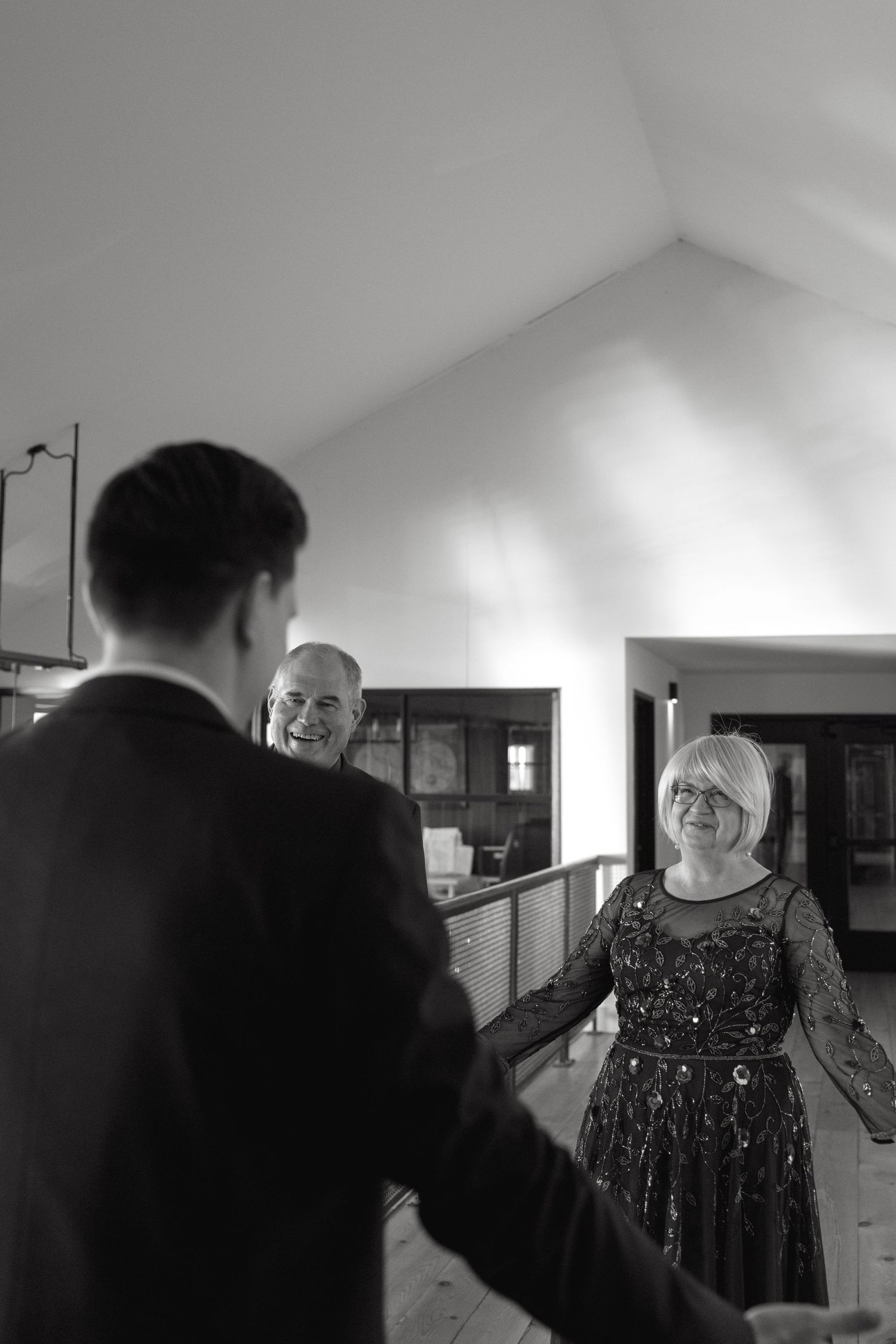 A black and white photo showing a woman in a sparkly dress greeting someone in an indoor hallway.