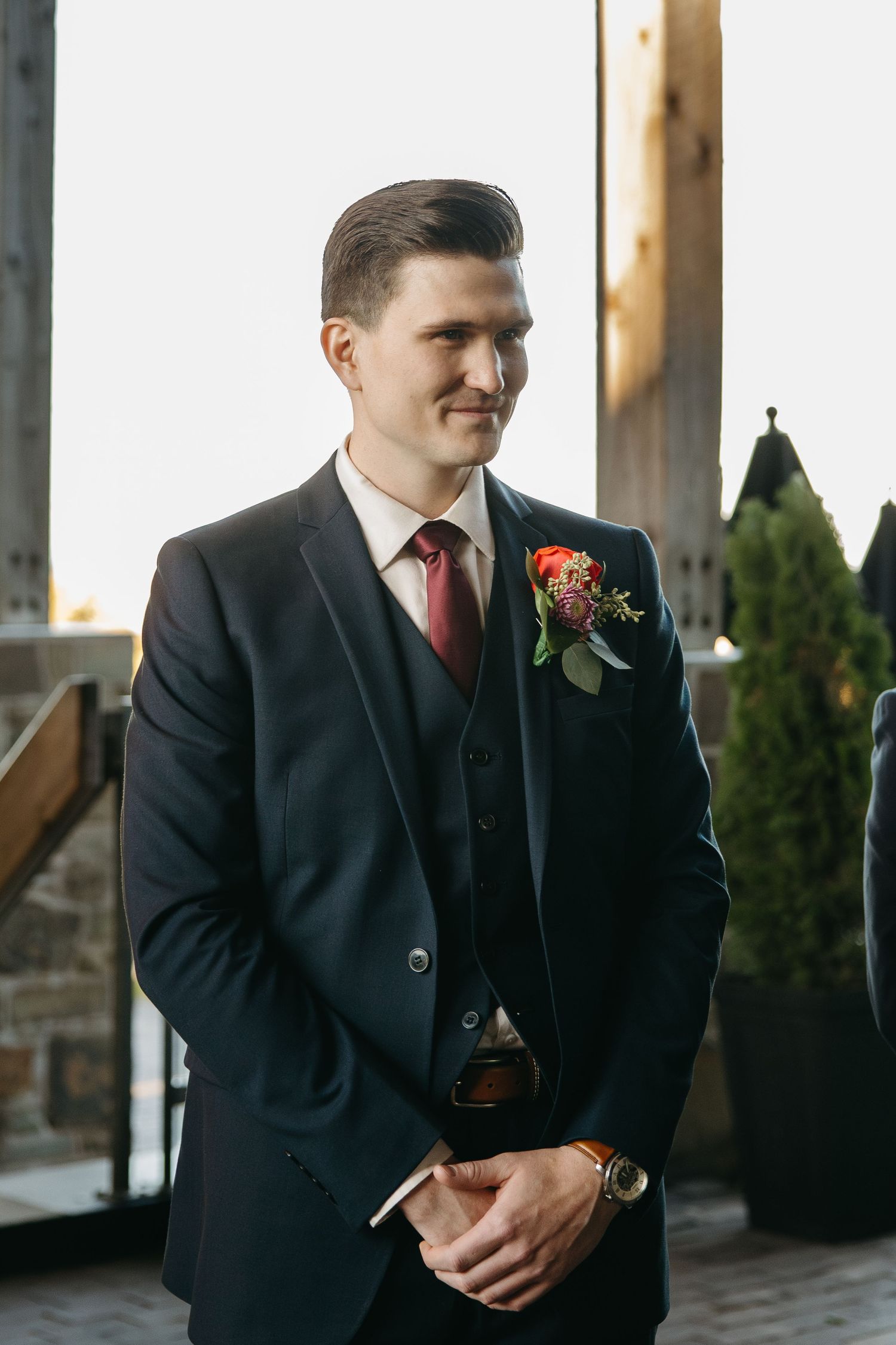 Groom in black suit and red tie standing in formal portraits before wedding ceremony.
