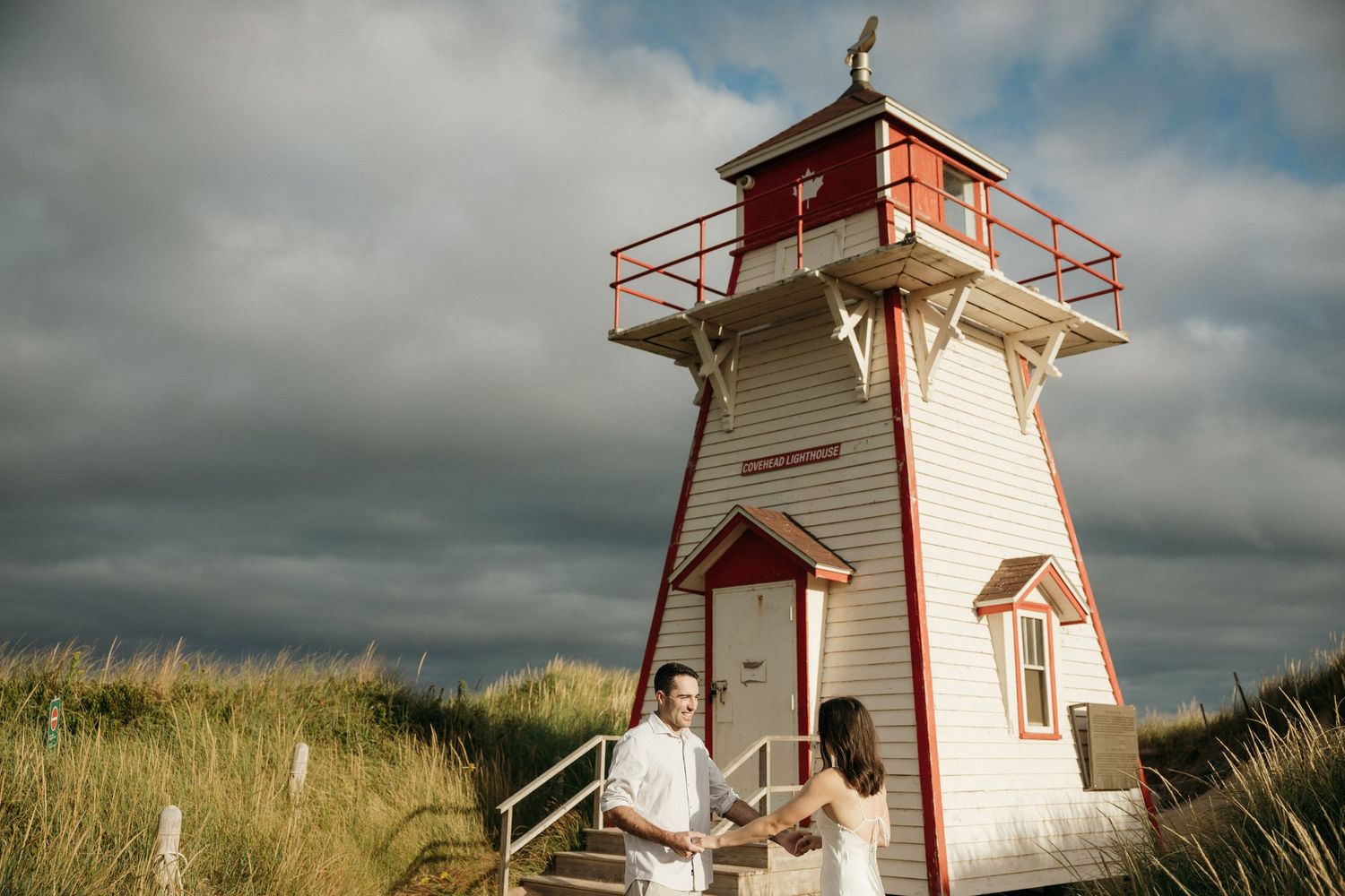 A white lighthouse with red trim stands against dramatic cloudy skies on a sandy beach.