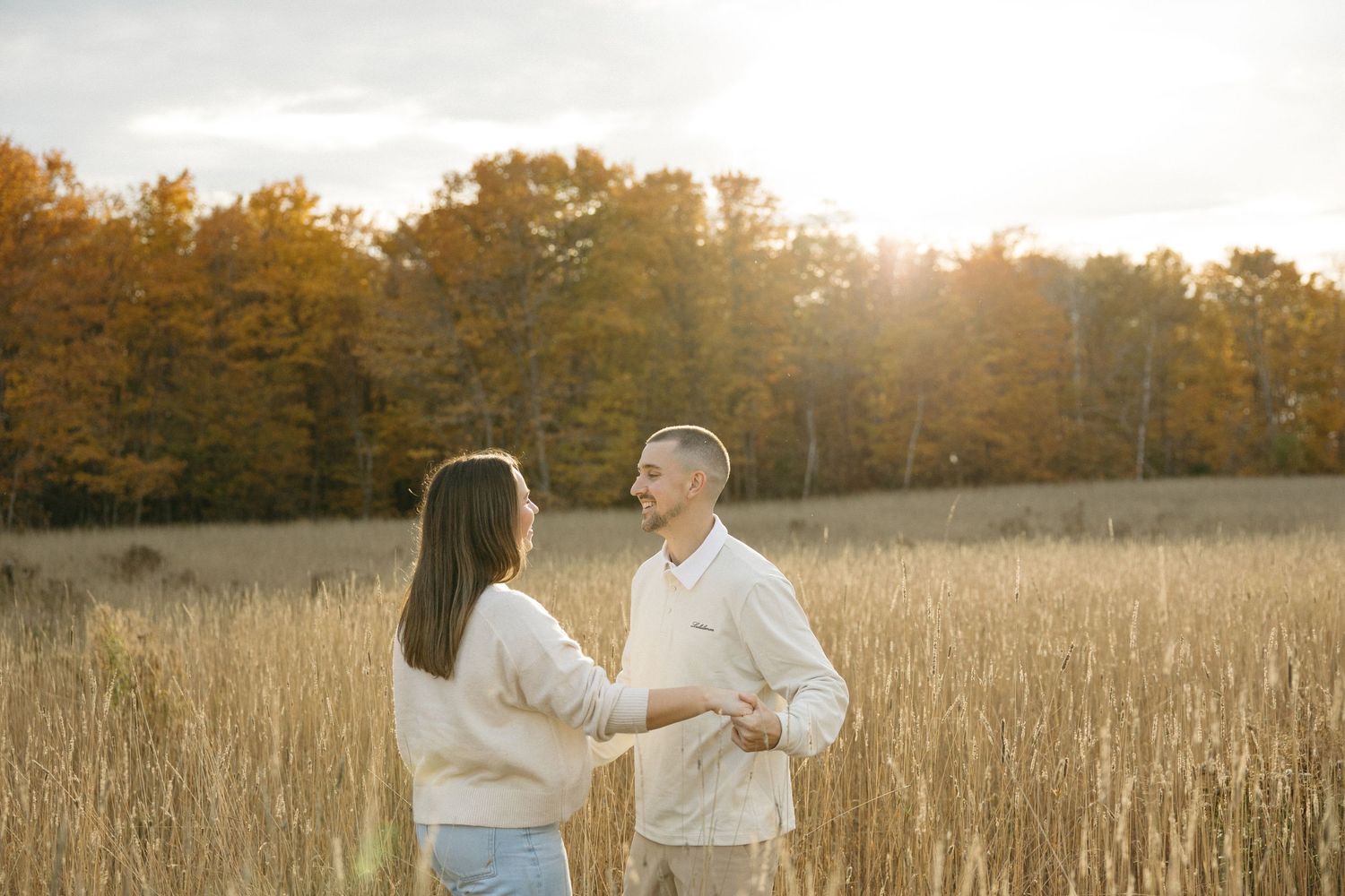 A couple in white outfits stands in a sunlit autumn field.