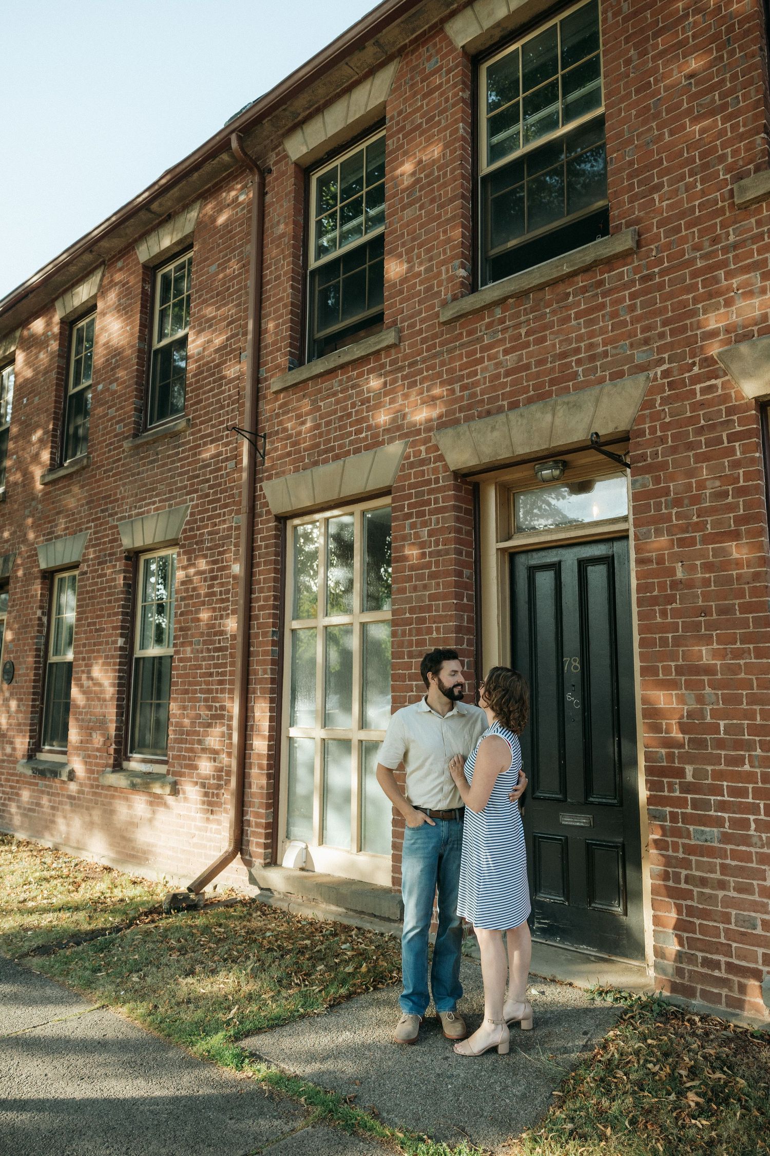 A couple poses together against a rustic brick building with black doors and windows.