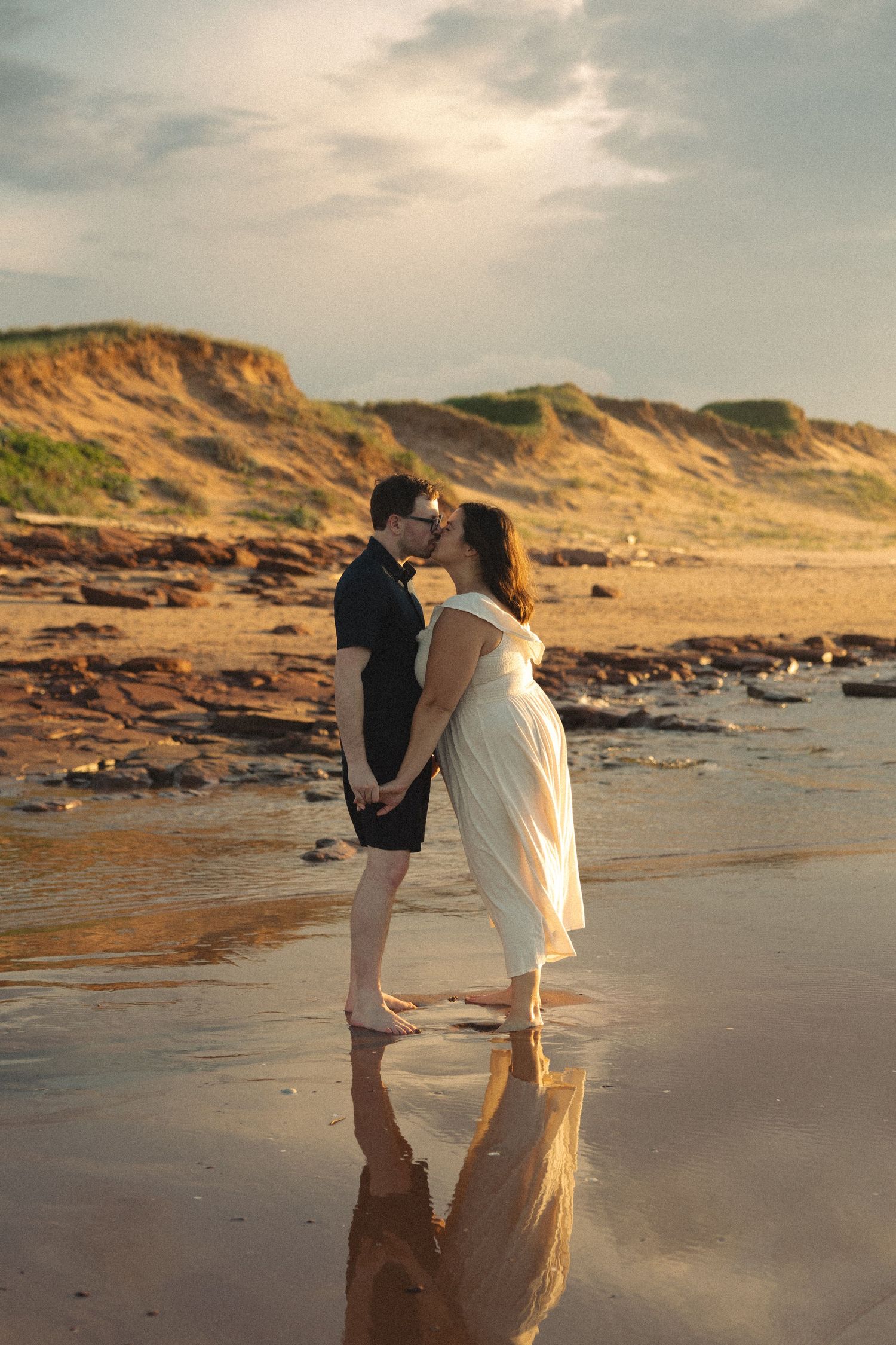 A couple shares a tender kiss while standing barefoot on a reflective beach at golden hour sunset.