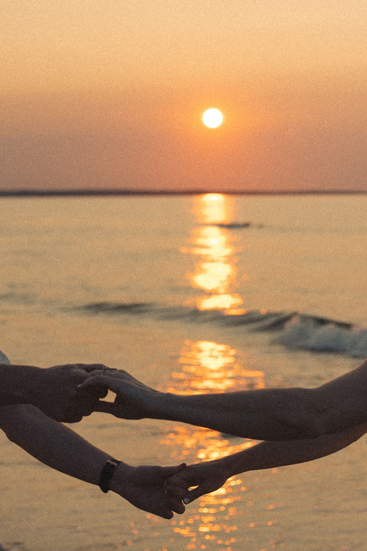 Two people holding hands at a beautiful golden ocean sunset, romantic beach scene with sun reflecting on calm water.