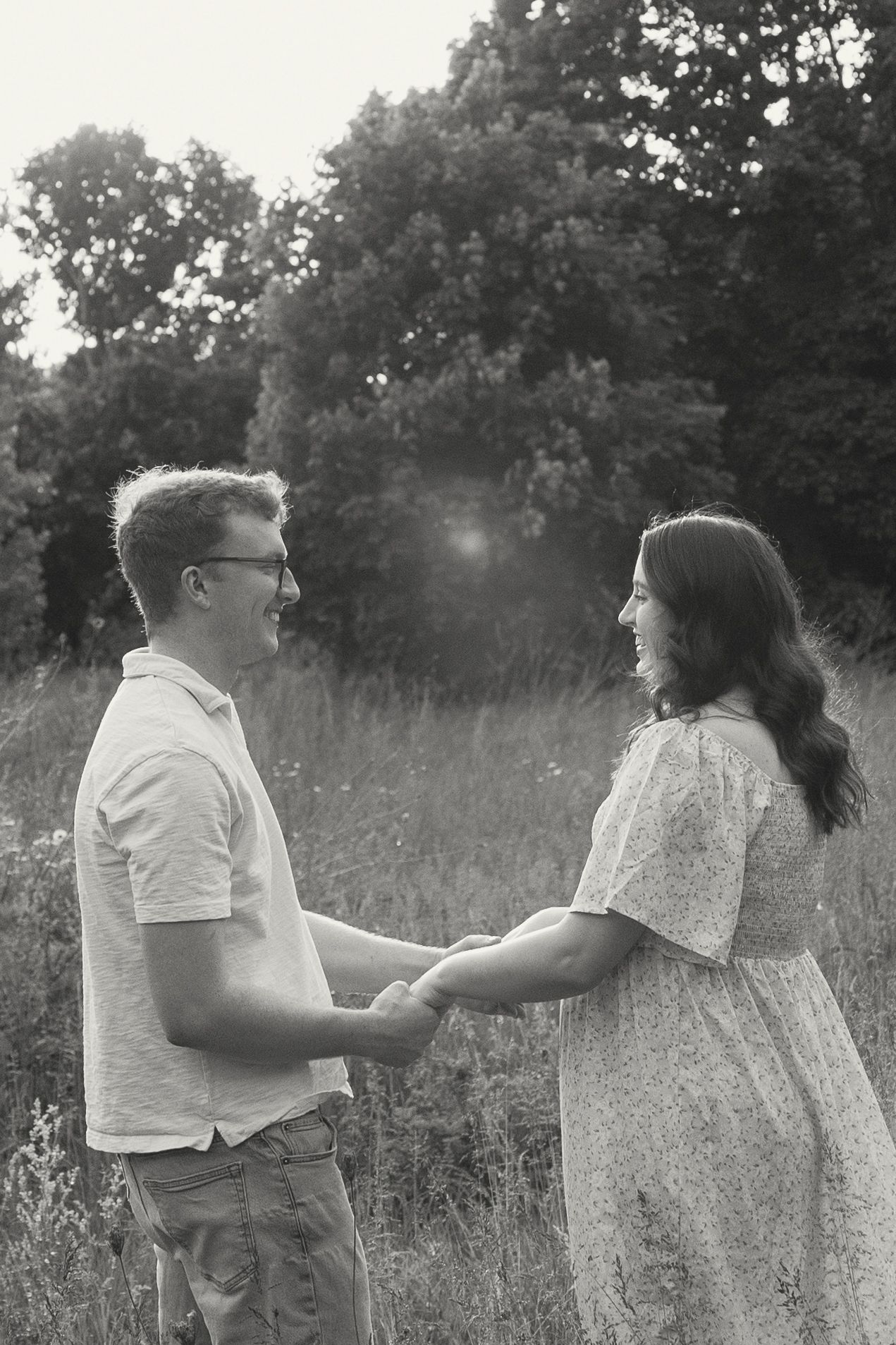 A couple shares a playful moment in a sunlit meadow, dancing and laughing together in black and white.