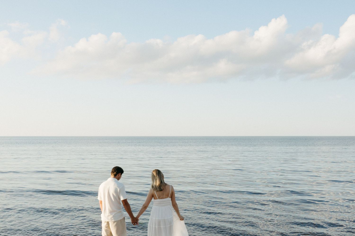 Couple holding hands facing the ocean on a serene beach, dressed in white wedding attire under a blue sky.