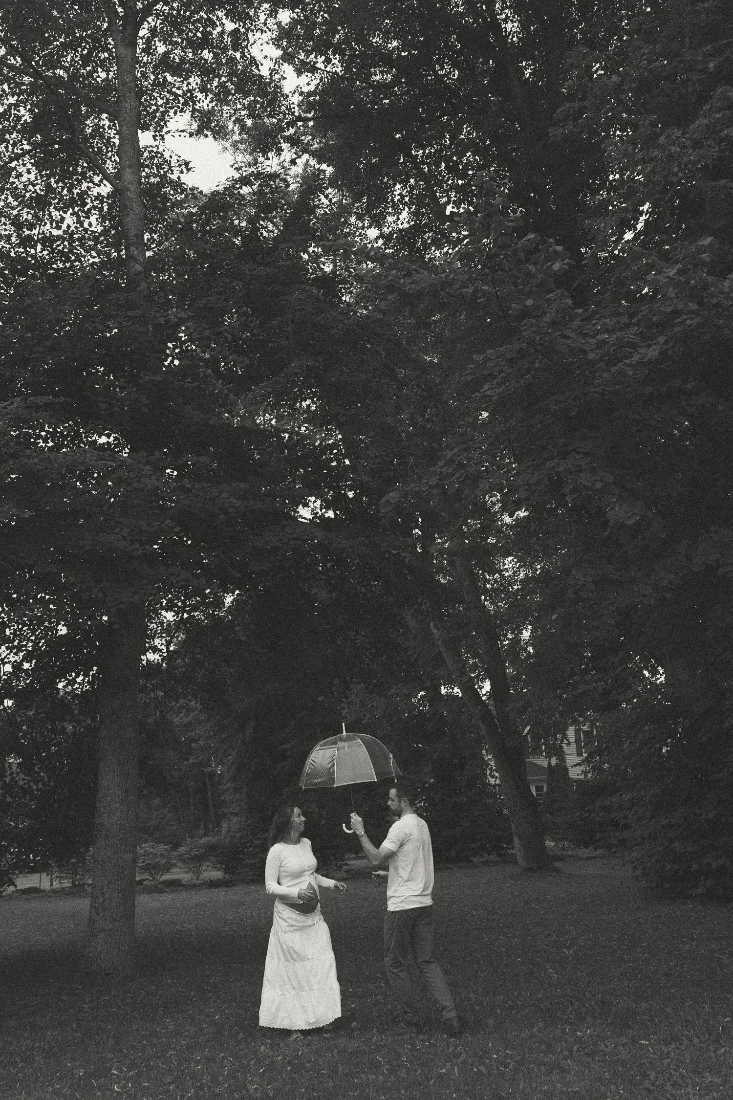 Black and white photo of a pregnant couple standing together under trees in a park, man photographing woman.