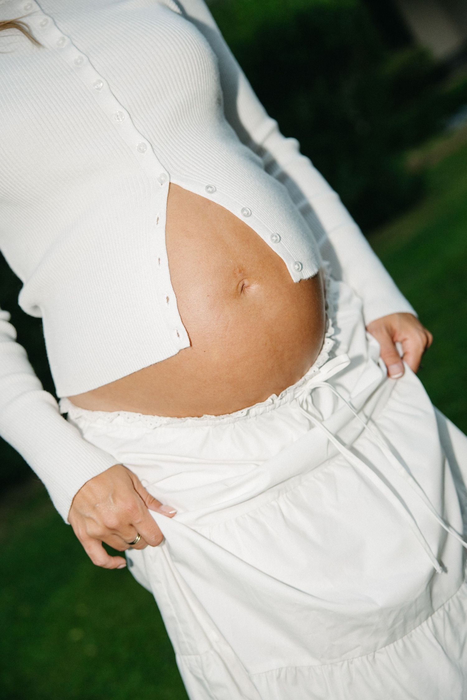 Pregnant woman in white ribbed top and skirt showing baby bump outdoors.
