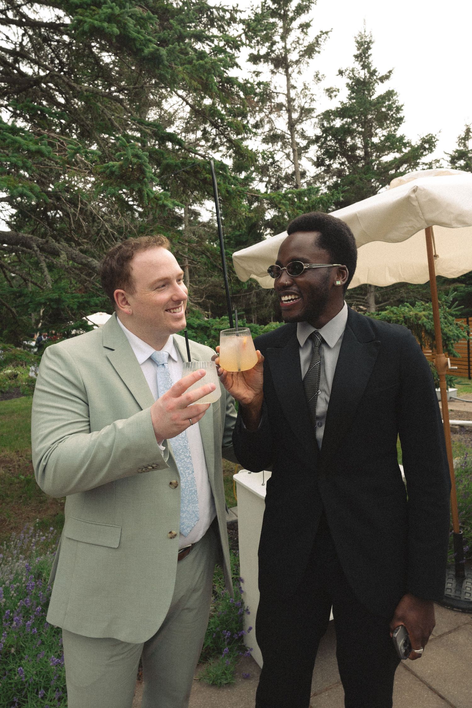 Two well-dressed wedding guests smile and toast with cocktails outdoors among pine trees.