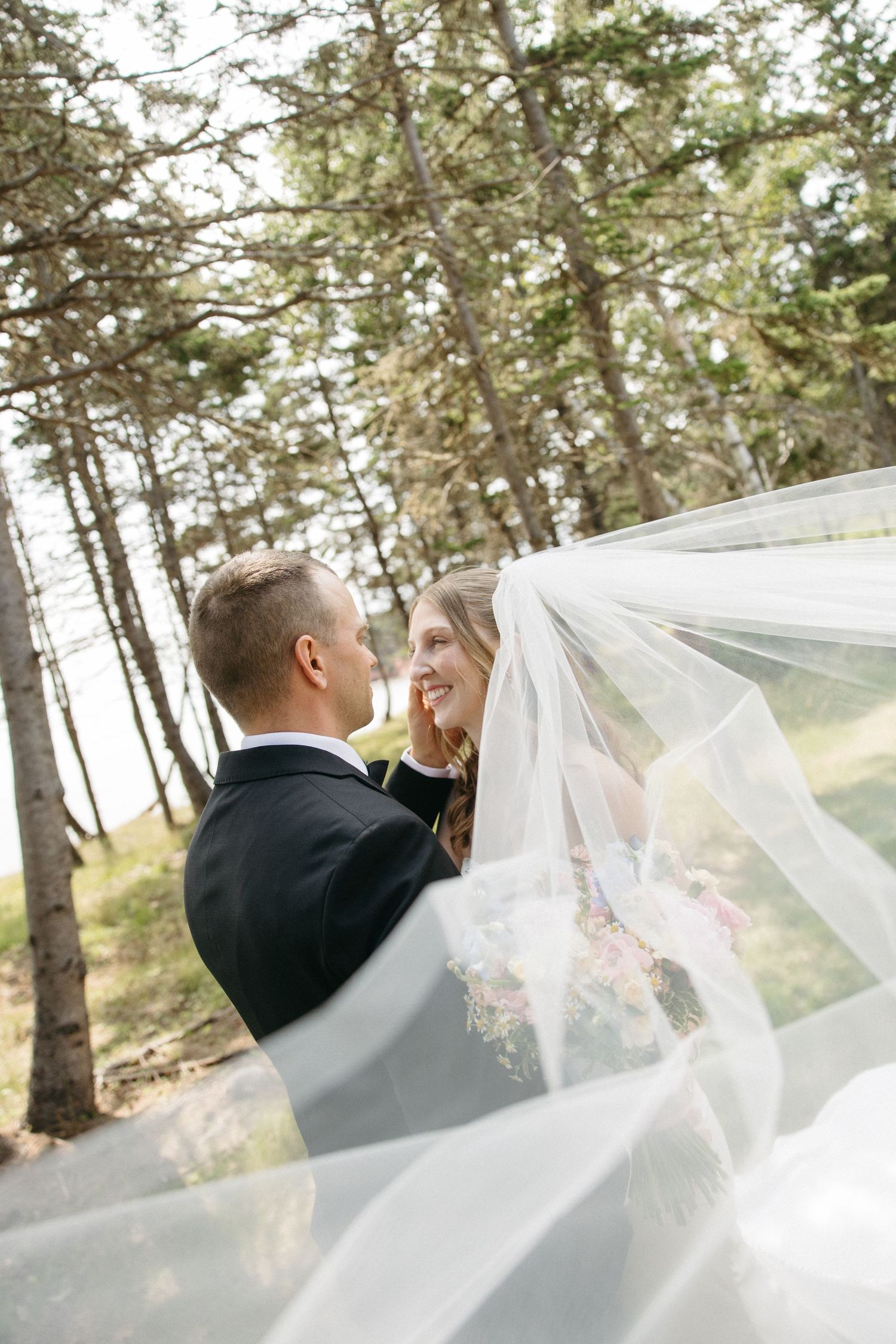 Bride and groom share a joyful moment under the flowing wedding veil in a forest setting.