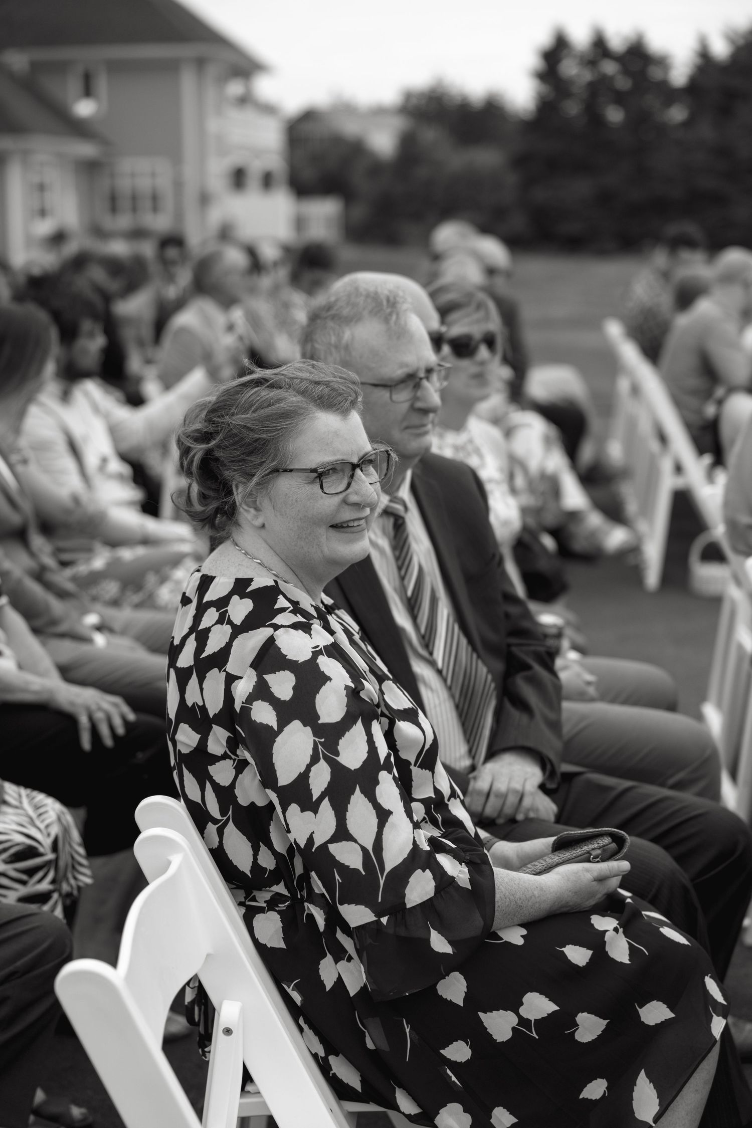Black and white photo of wedding guests seated in white chairs smiling during an outdoor ceremony.
