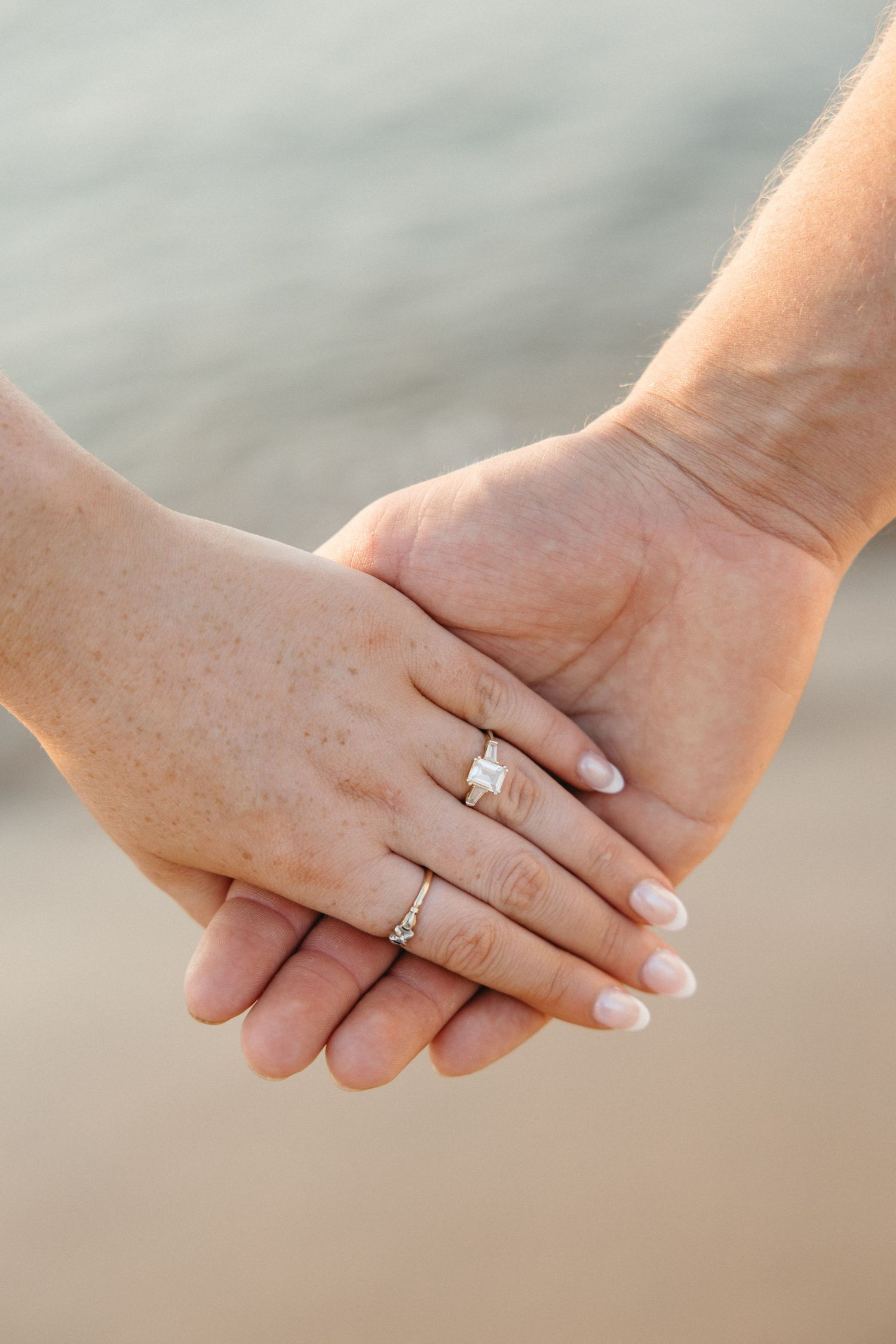 Close-up of couple holding hands on beach, woman wearing elegant diamond engagement ring and wedding band.