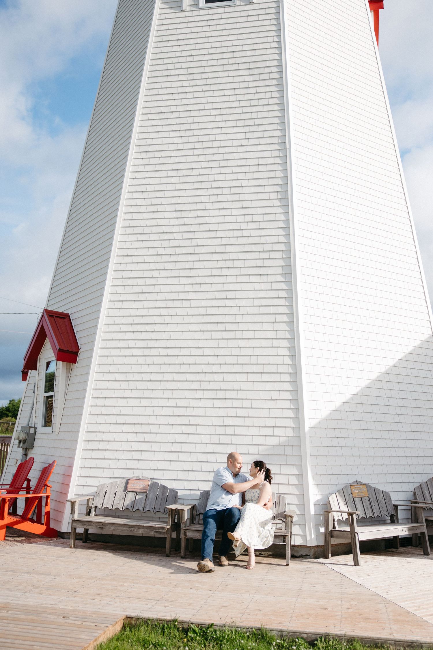 Couple sitting together on wooden bench beside white lighthouse on sunny day.