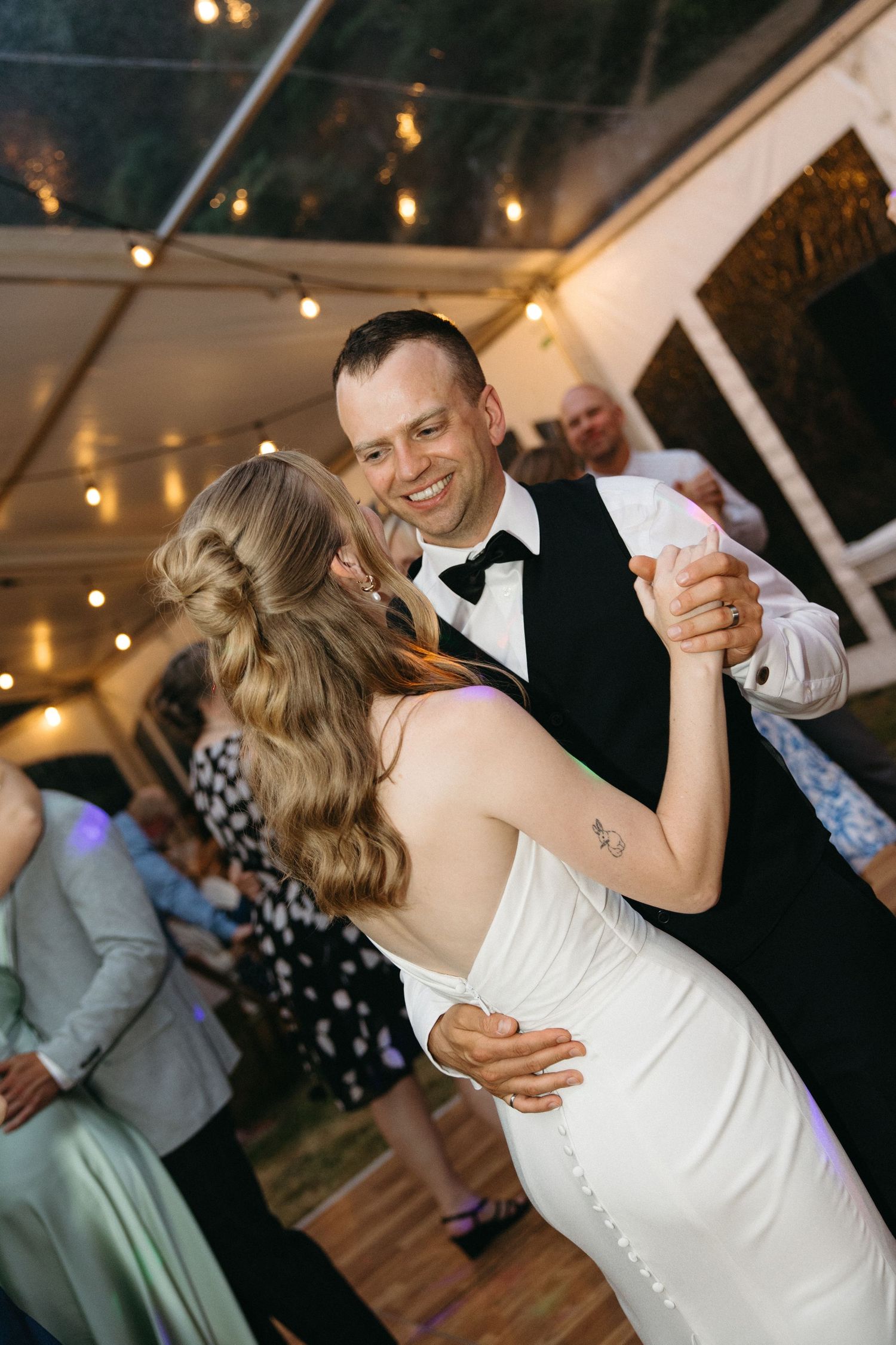 A smiling groom in a black vest and bow tie shares a first dance with his bride in a white gown at a tented wedding reception.