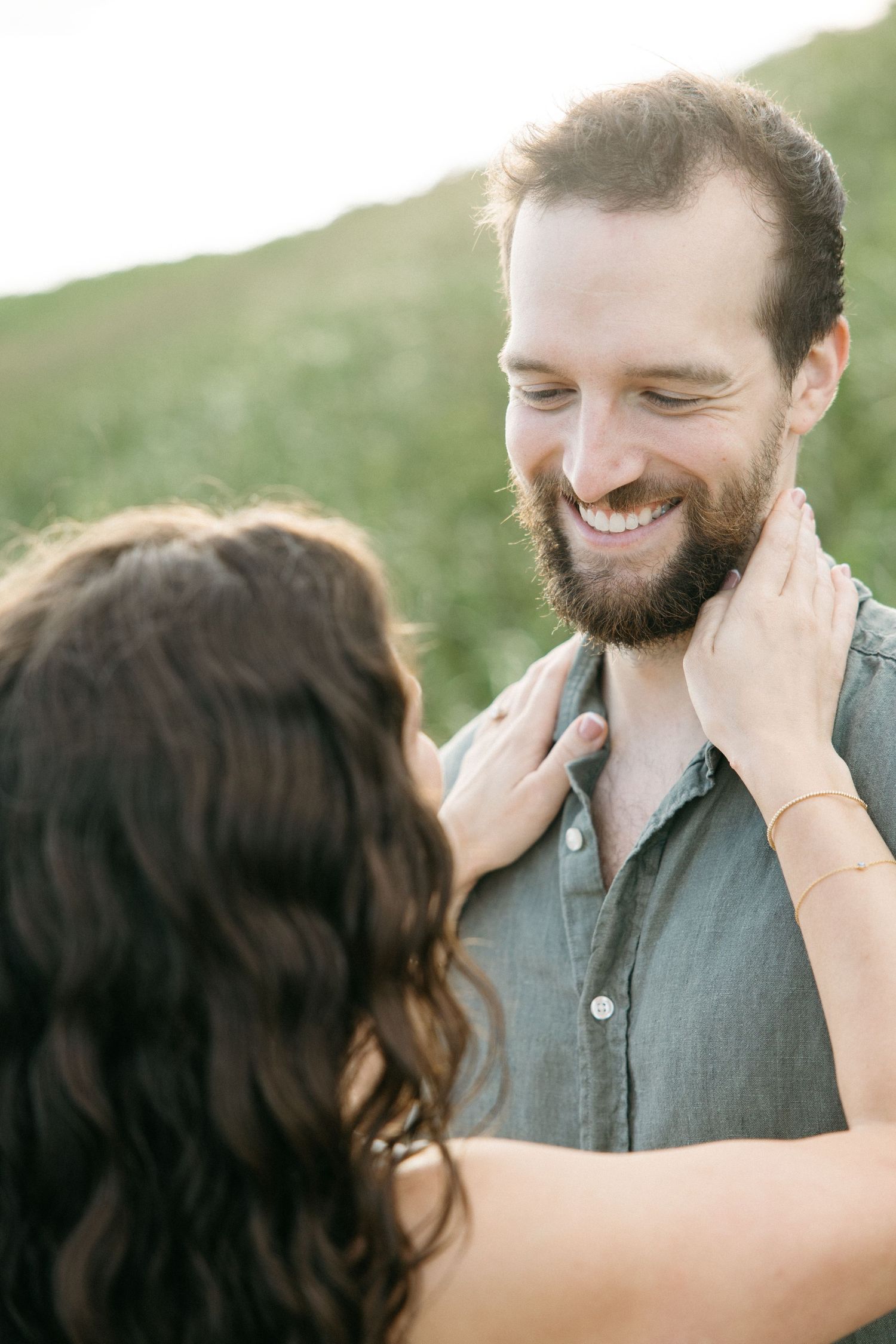 Smiling bearded man in gray shirt being tenderly held by a woman outdoors in a green field.