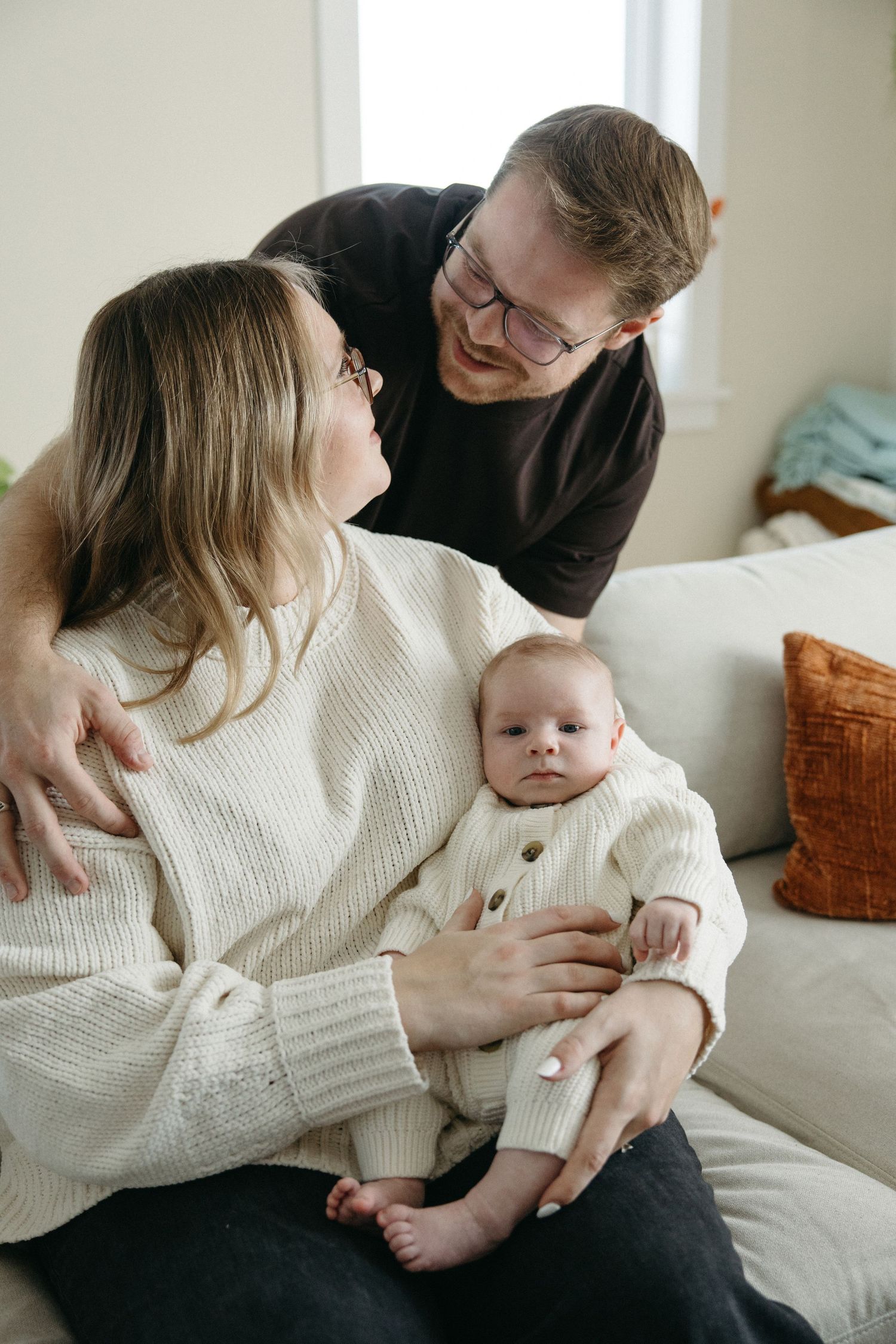 Intimate family photos show parents in casual attire sharing sweet moments with their infant on a light colored sofa.