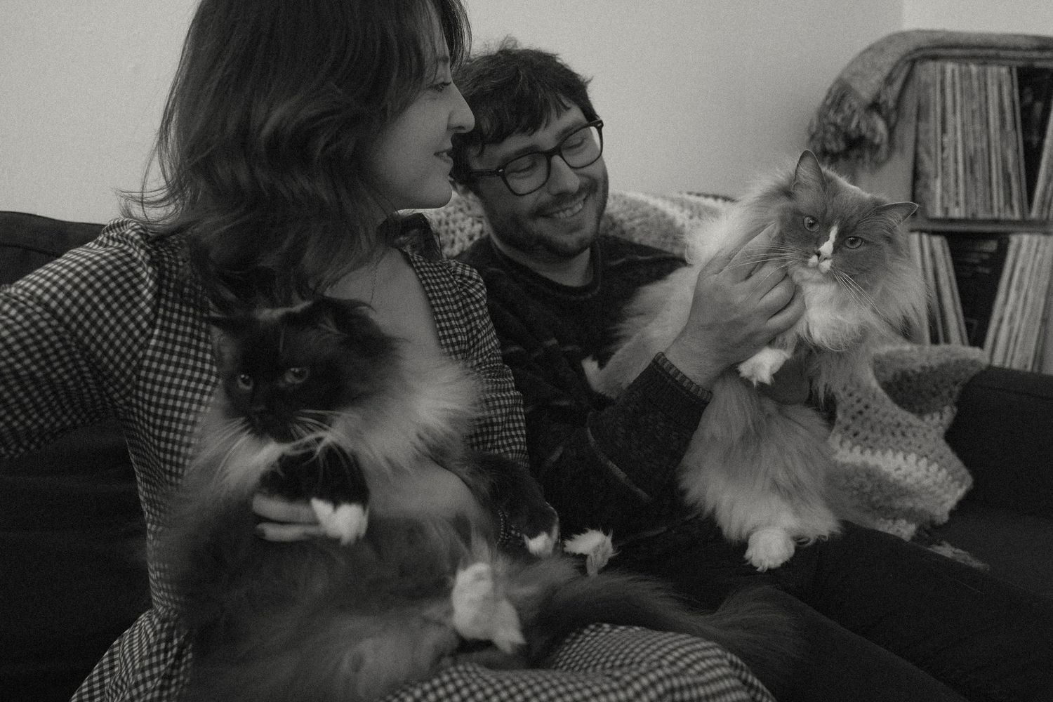 Black and white photo of a couple sitting on a couch, each holding a fluffy cat, with bookshelves in the background.