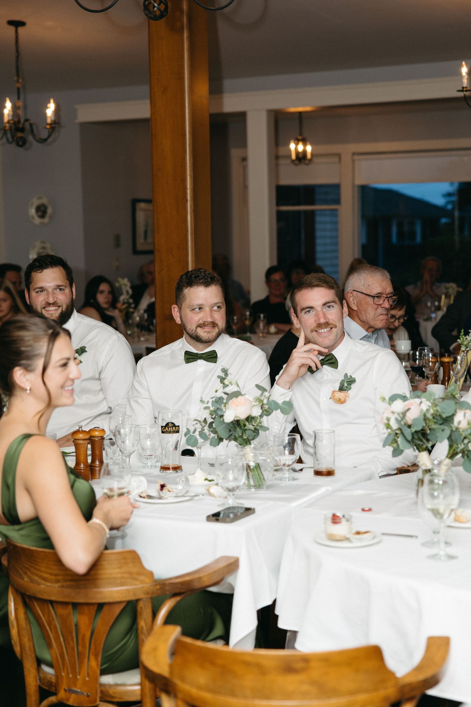 Wedding guests in white shirts and green bow ties smiling at reception dinner tables with floral centerpieces.