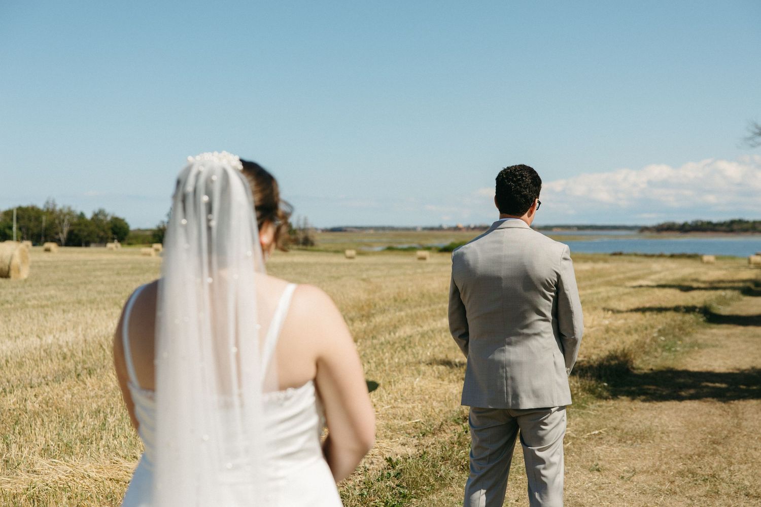 Bride in white gown and veil approaching groom in gray suit standing before a scenic coastal field and blue sky.